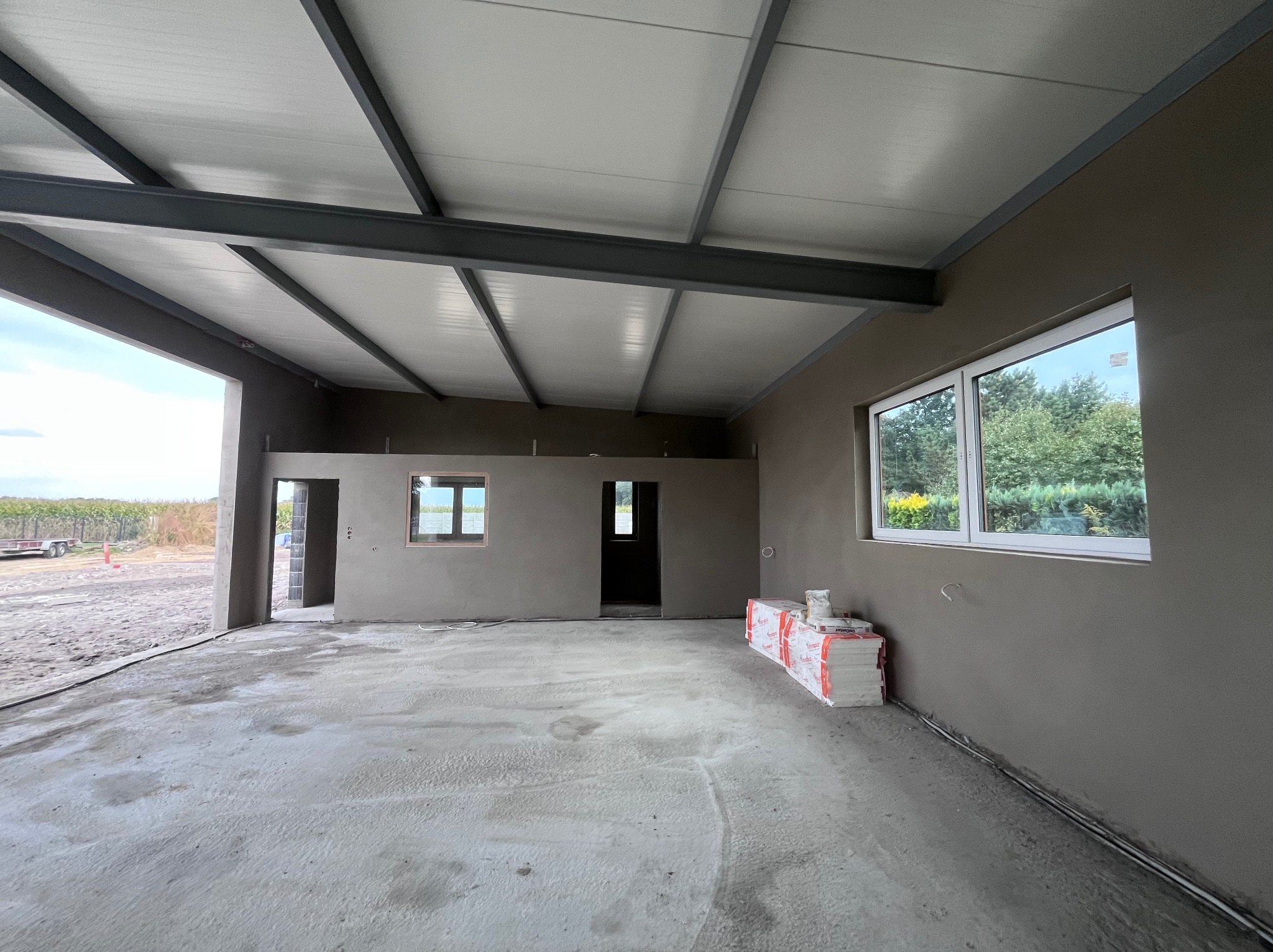 Wnętrze w stanie surowym: szare tynki, betonowa podłoga, białe okna, metalowa konstrukcja dachu. Materiały budowlane w rogu pomieszczenia.