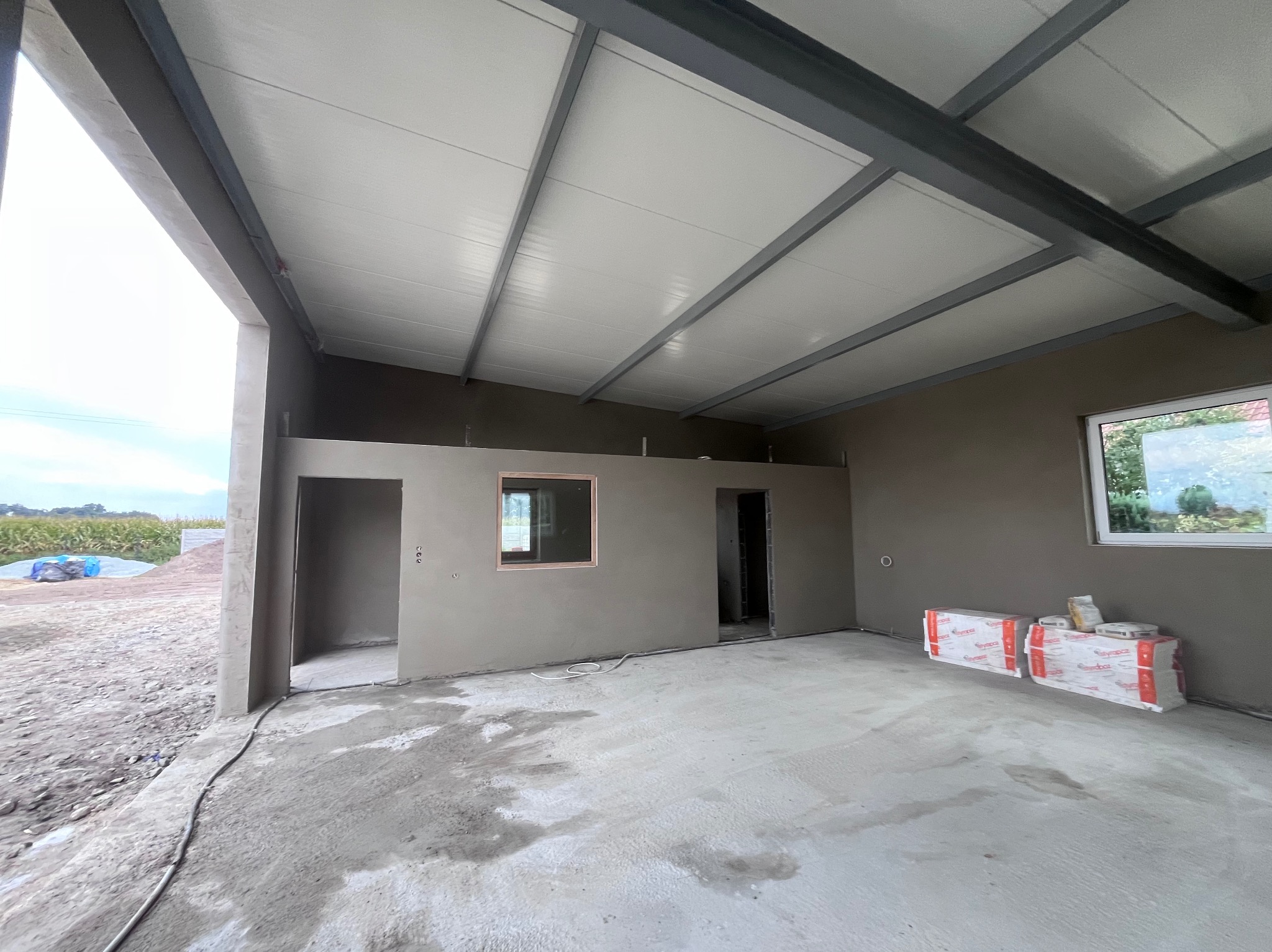 Wnętrze w stanie surowym: ściany po tynkowaniu, widoczne okno, sufit z metalową konstrukcją, podłoga betonowa. Na ścianie leżą zapakowane materiały budowlane.