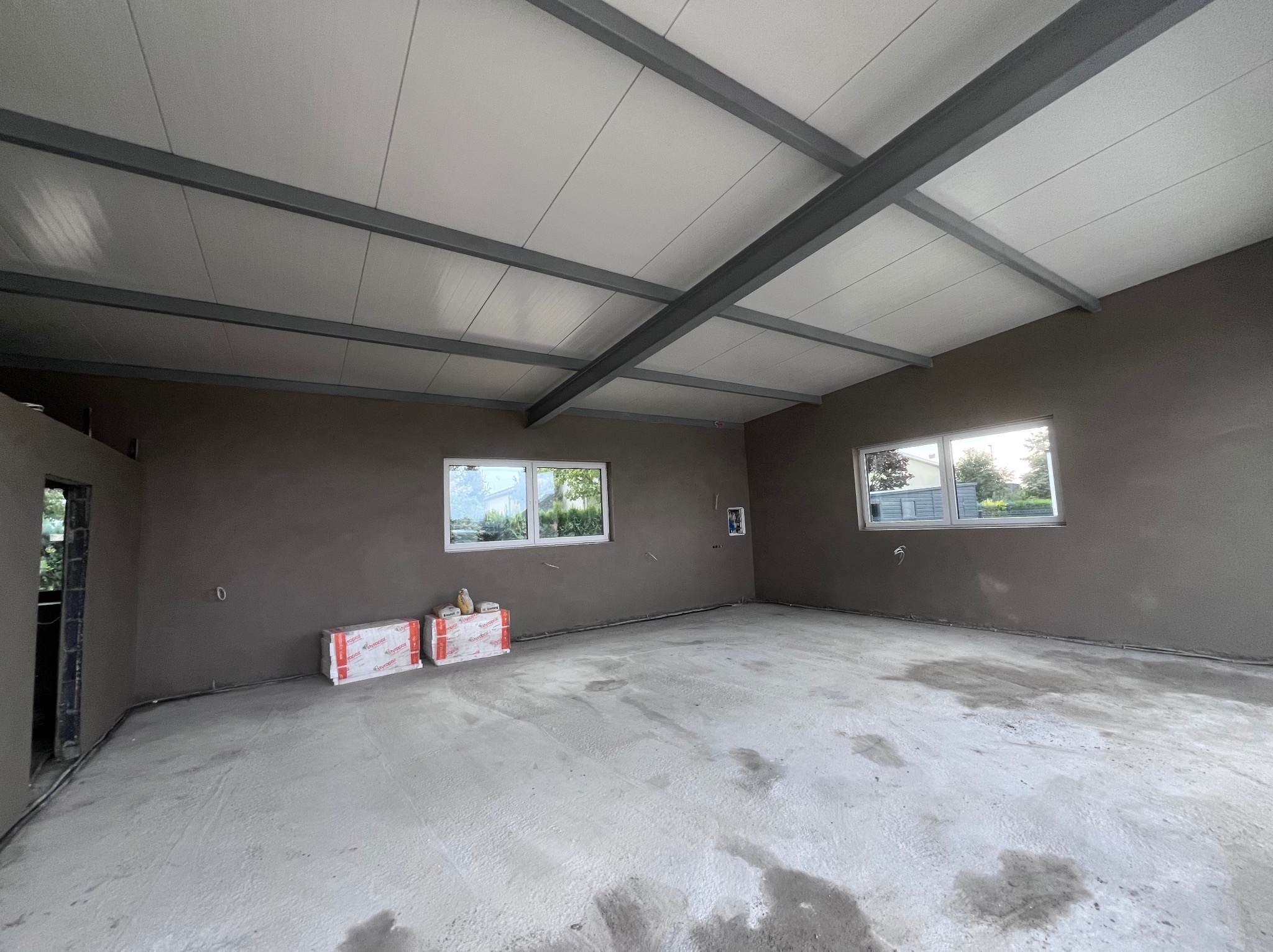 Wnętrze w stanie surowym: surowe ściany w odcieniach szarości, betonowa podłoga i biały sufit z widoczną konstrukcją nośną. Dwa okna wpuszczają naturalne światło.