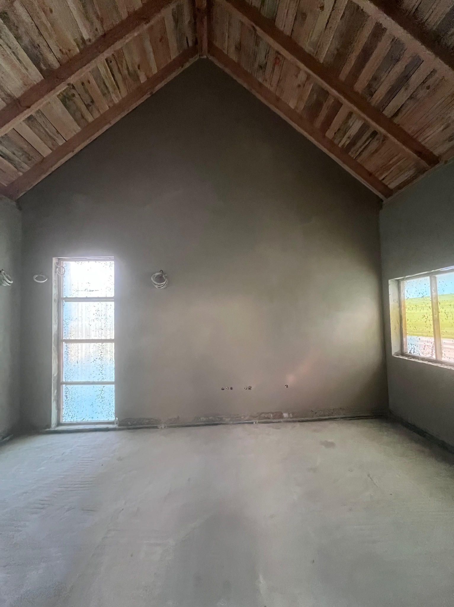 Surowe wnętrze po tynkowaniu, z drewnianym sufitem w kształcie litery A, widoczne okna i szare ściany. Wnętrze w trakcie prac wykończeniowych.