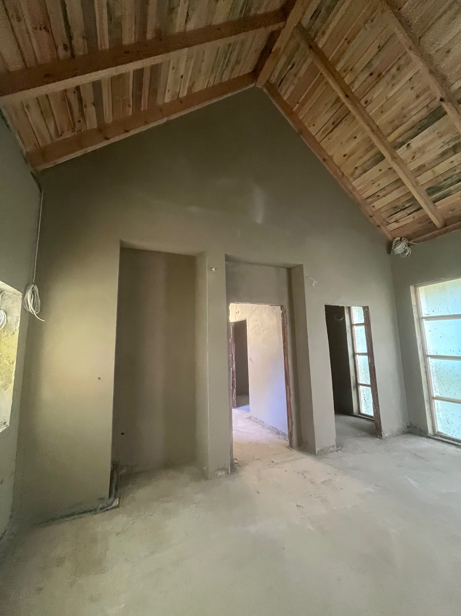 Wnętrze z tynkami w kolorze szarości, surowy drewniany strop, widoczne otwory drzwiowe i okienne, podłoga w trakcie prac wykończeniowych.