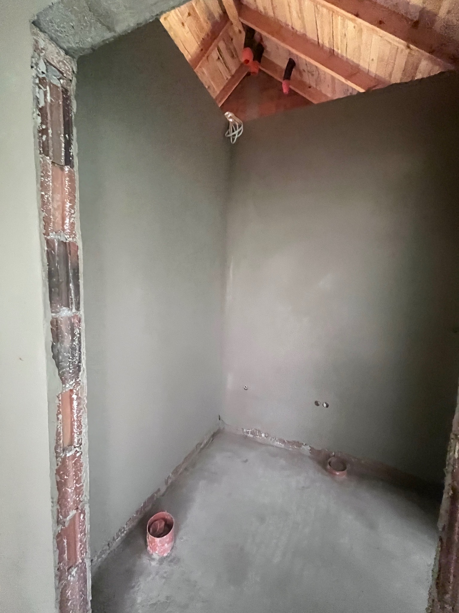 Wnętrze pomieszczenia po nałożeniu tynku, widoczne fragmenty cegieł przy ościeżnicy. Poddasze z drewnianą konstrukcją i wystającymi elementami instalacji.