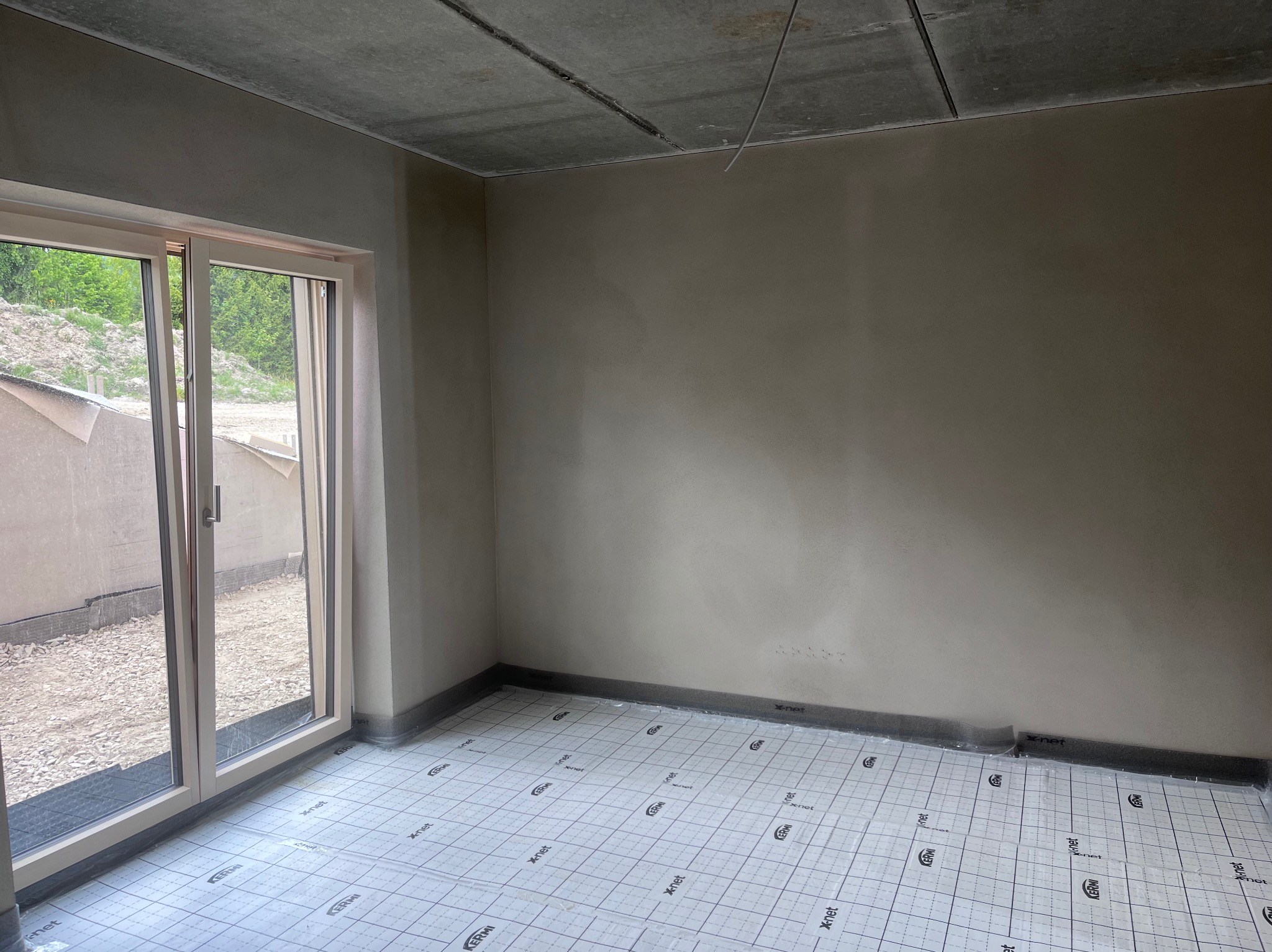 Pokój po tynkowaniu ścian, z oknem balkonowym i przygotowaną podłogą z izolacją, gotowy do dalszych prac wykończeniowych. Widoczne surowe wykończenie sufitu.