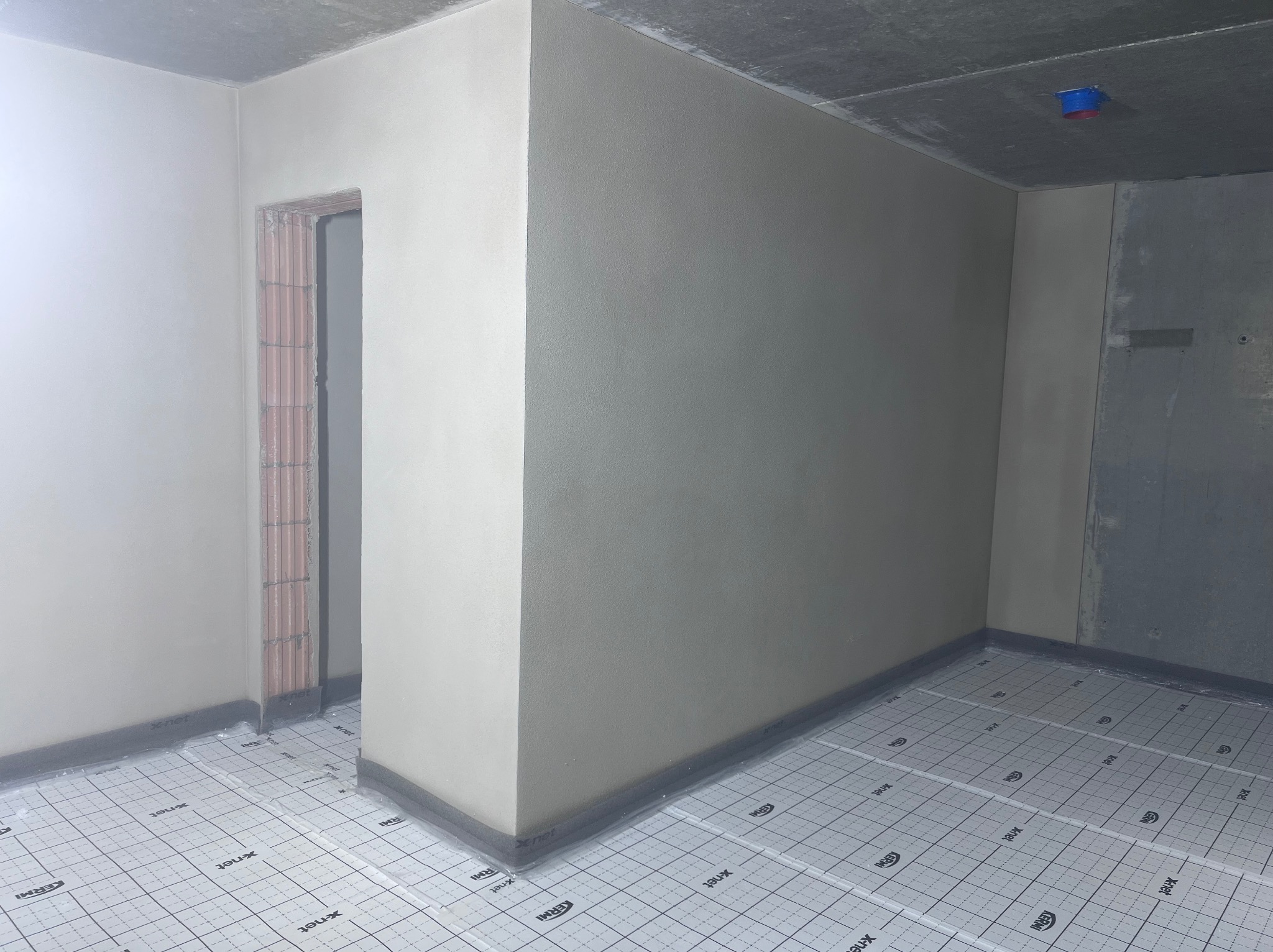 Wnętrze w trakcie prac wykończeniowych: świeżo otynkowane ściany, surowy betonowy sufit z instalacją i podłoga przygotowana pod ogrzewanie podłogowe.