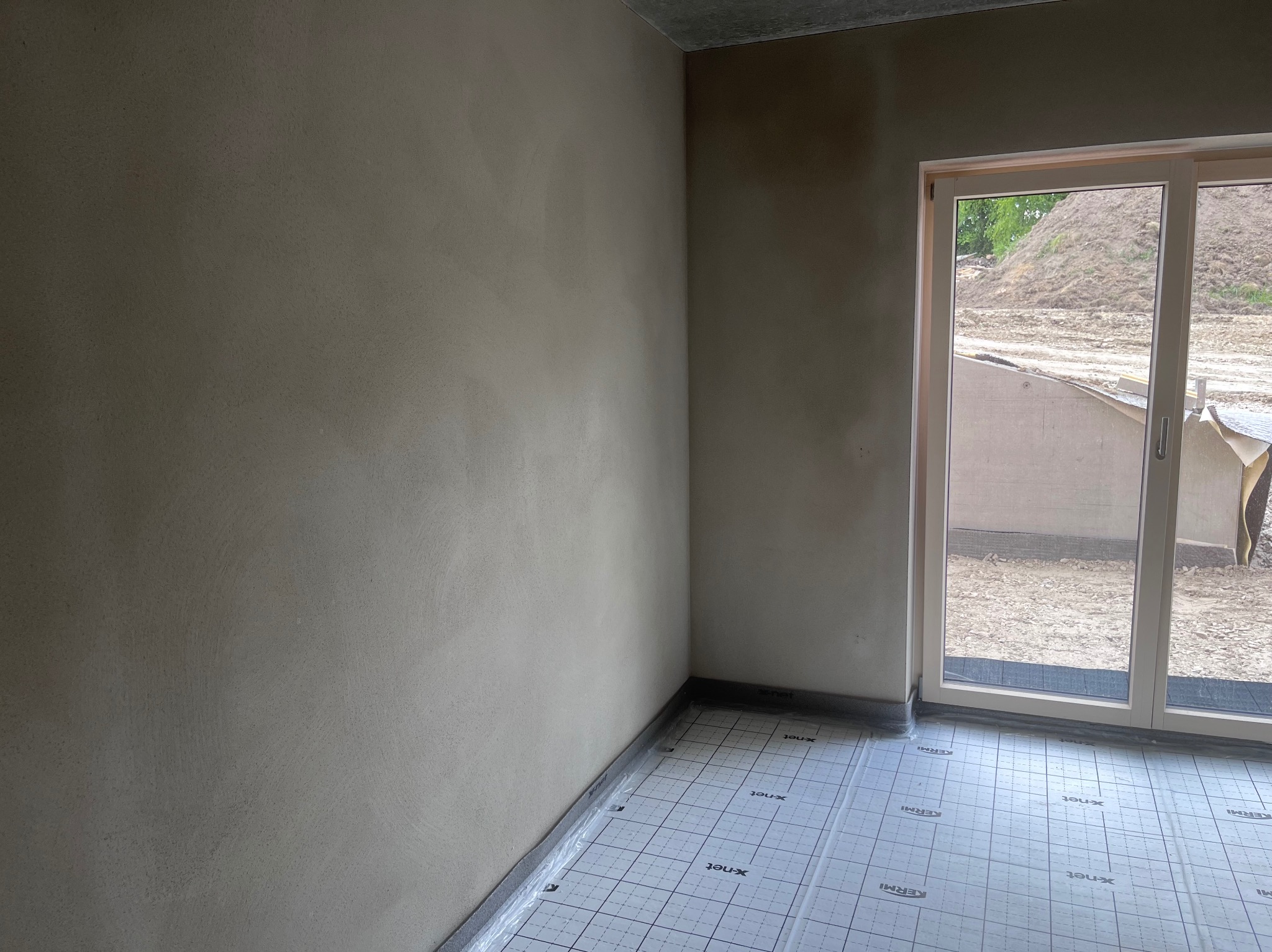 Ściana po tynkowaniu z widoczną siatką pod ogrzewanie podłogowe i oknem z widokiem na zewnątrz. Surowy betonowy sufit i widoczne elementy wykończeniowe.