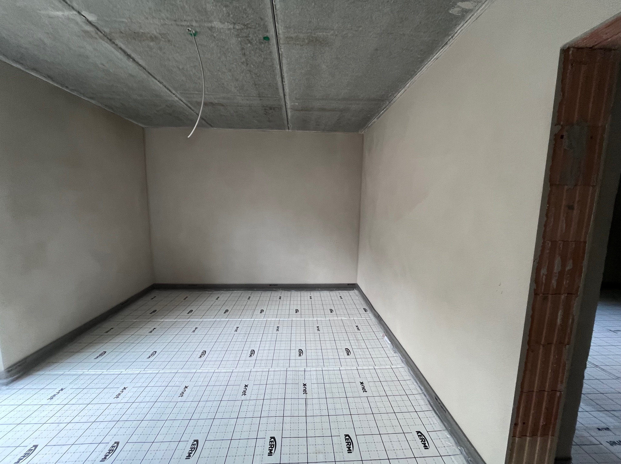 Surowe wnętrze z przygotowanym podłożem pod ogrzewanie podłogowe, ściany po szpachlowaniu. Na suficie widoczne płyty kartonowo-gipsowe. W tle fragment muru.