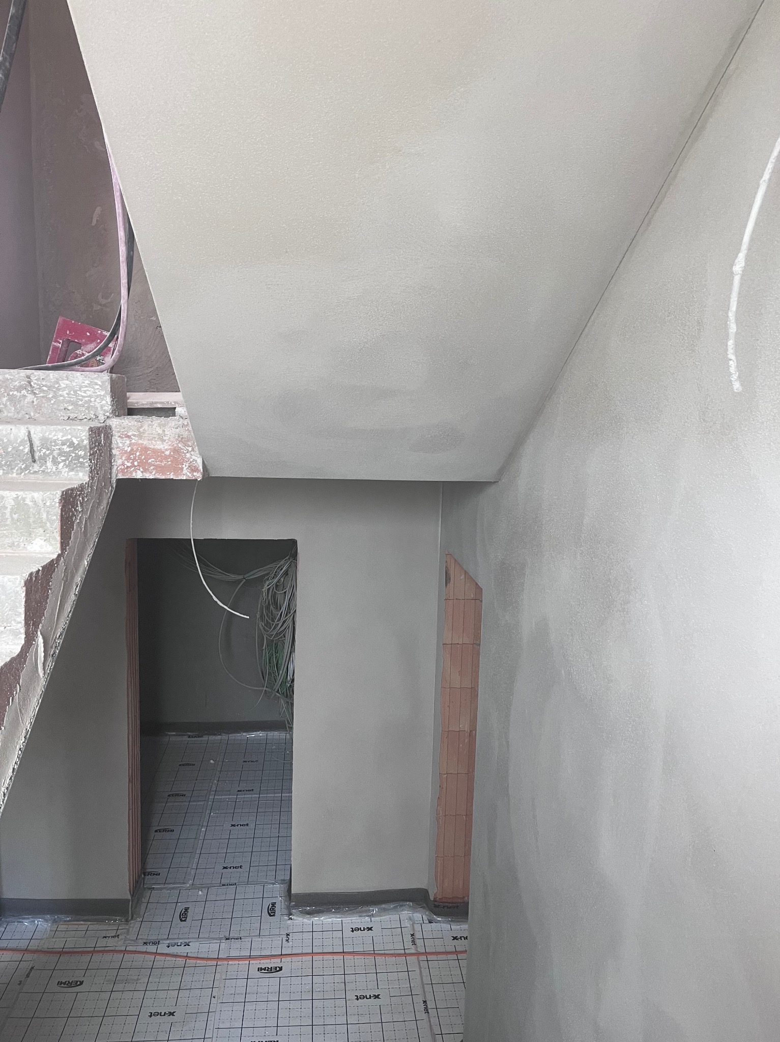 Wnętrze w trakcie prac: świeżo otynkowane ściany i sufit, widoczne schody w surowym stanie oraz instalacja elektryczna w otworze drzwiowym. Podłoga zabezpieczona.