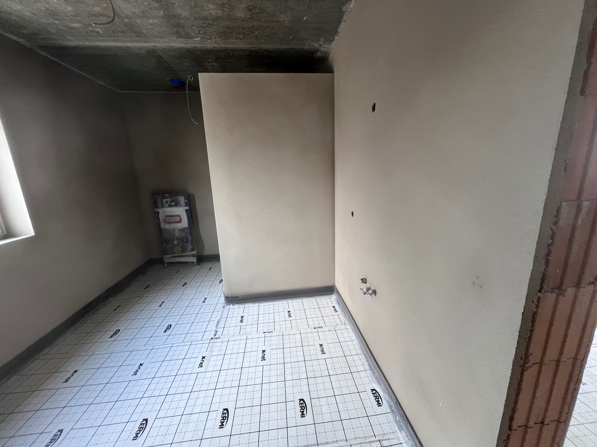 Wnętrze w trakcie prac wykończeniowych: ściany pokryte jasną gładzią, podłoga zabezpieczona folią ochronną z nadrukiem, widoczny narożnik z cegły.