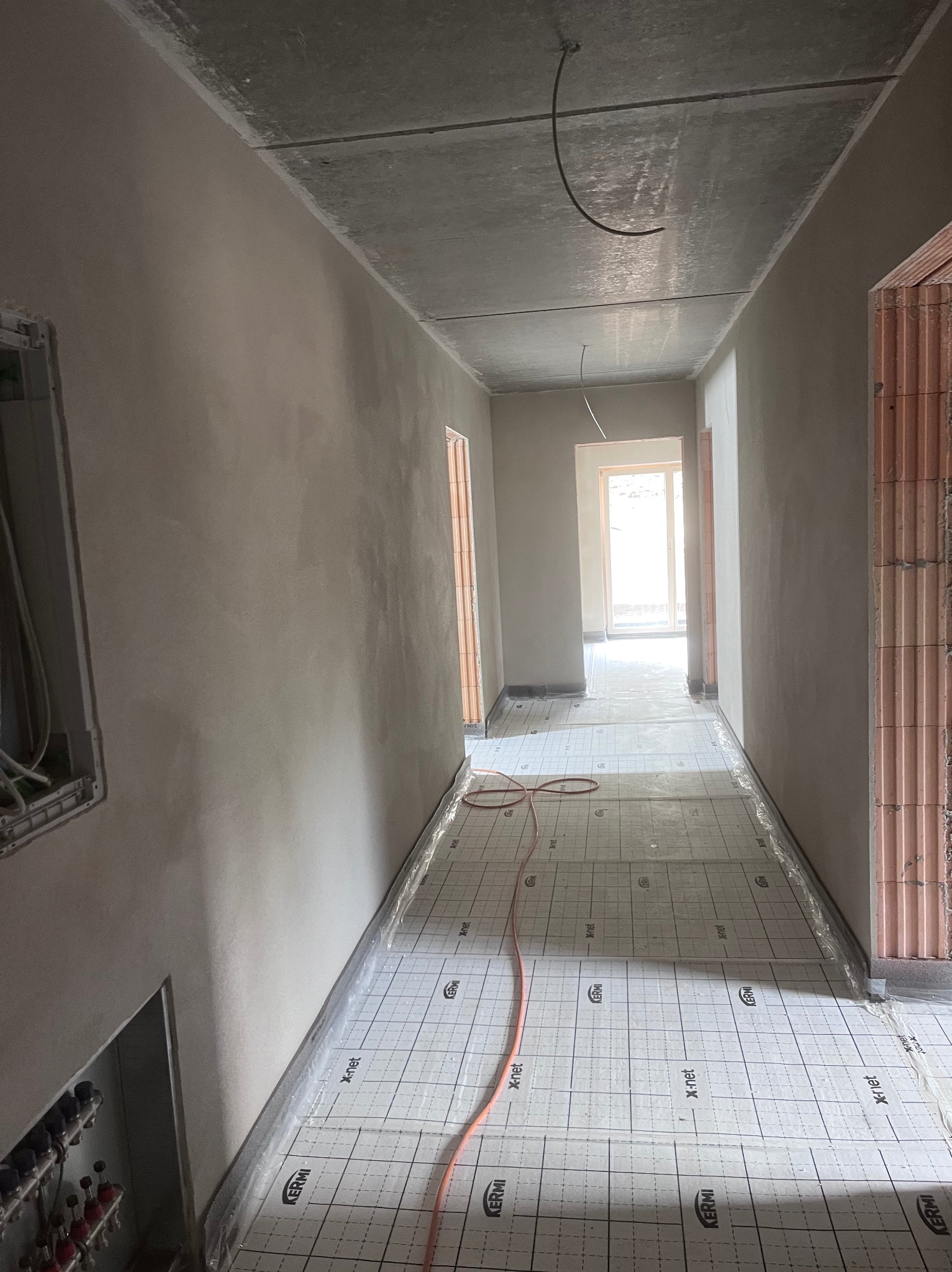 Korytarz w trakcie prac wykończeniowych: świeżo otynkowane ściany, instalacja ogrzewania podłogowego z rurkami na izolacji. Widoczne kable wystające z sufitu.