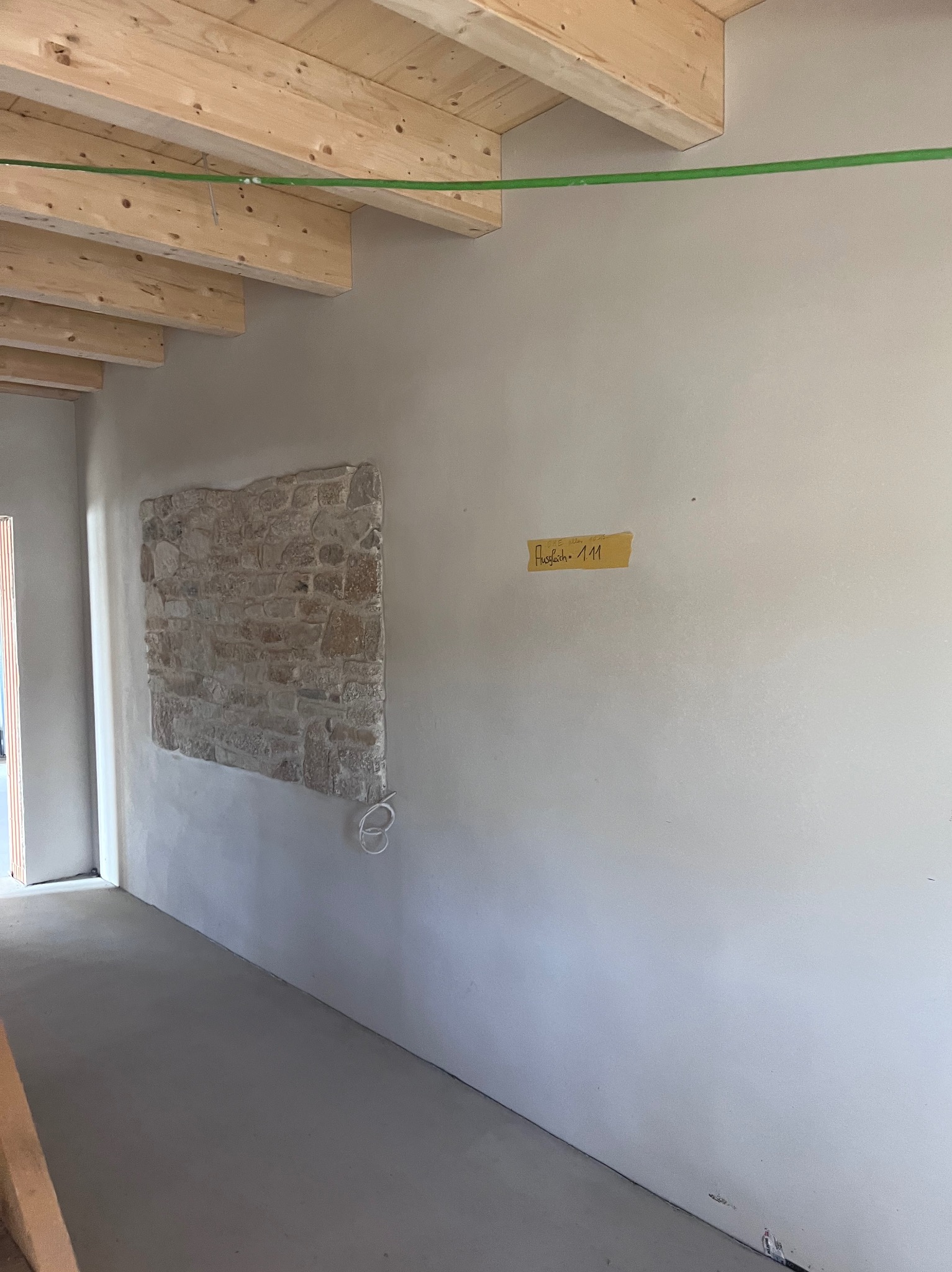 Wnętrze w trakcie prac wykończeniowych: fragment ściany z dekoracyjnym elementem z kamienia, świeżo wylana posadzka, drewniane belki stropowe, żółta naklejka na ścianie.