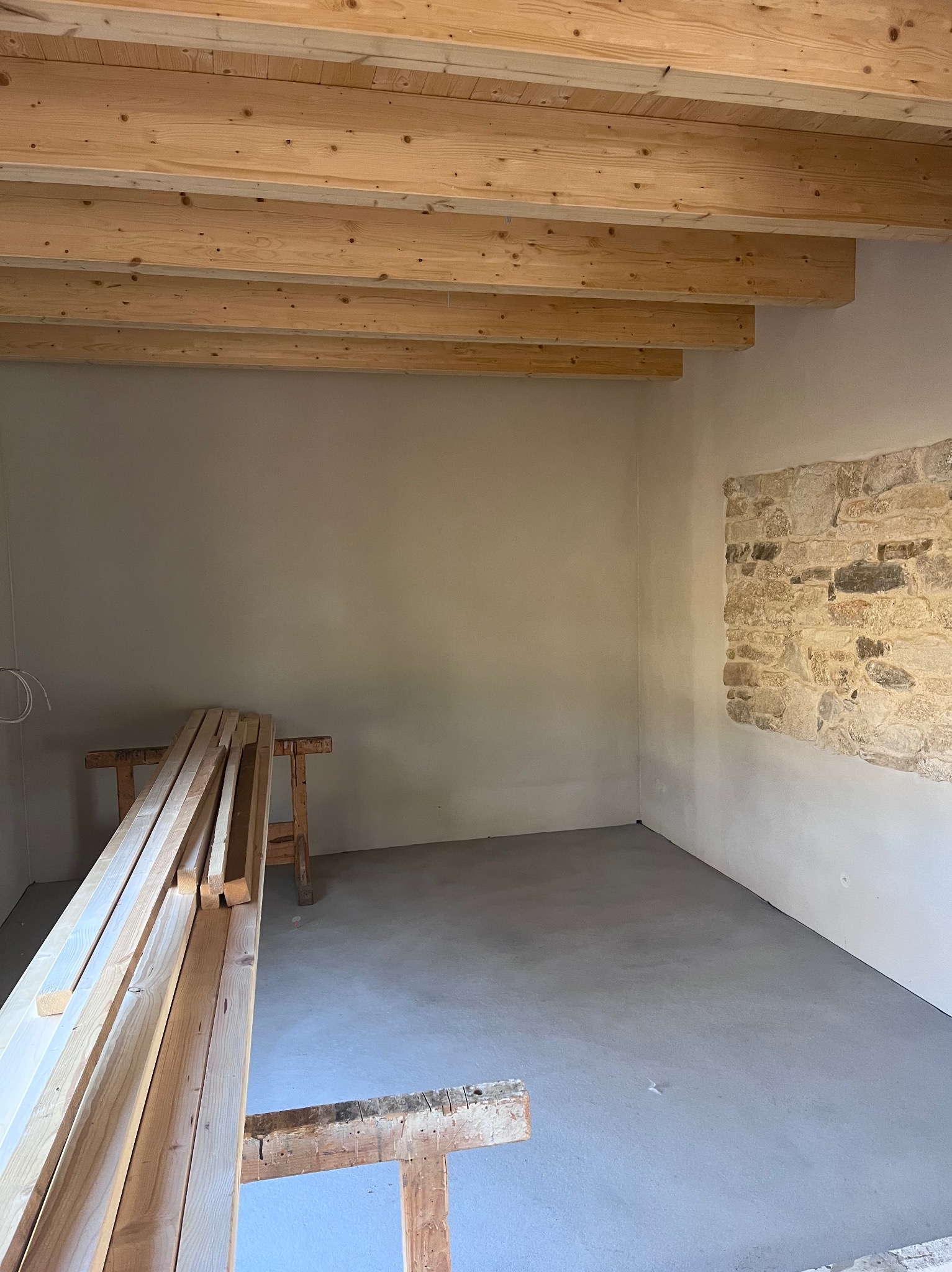 Surowe drewno na tle świeżo wylanej, szarej posadzki w pomieszczeniu z odsłoniętą kamienną ścianą i drewnianą konstrukcją stropu. Wnętrze w trakcie prac wykończeniowych.