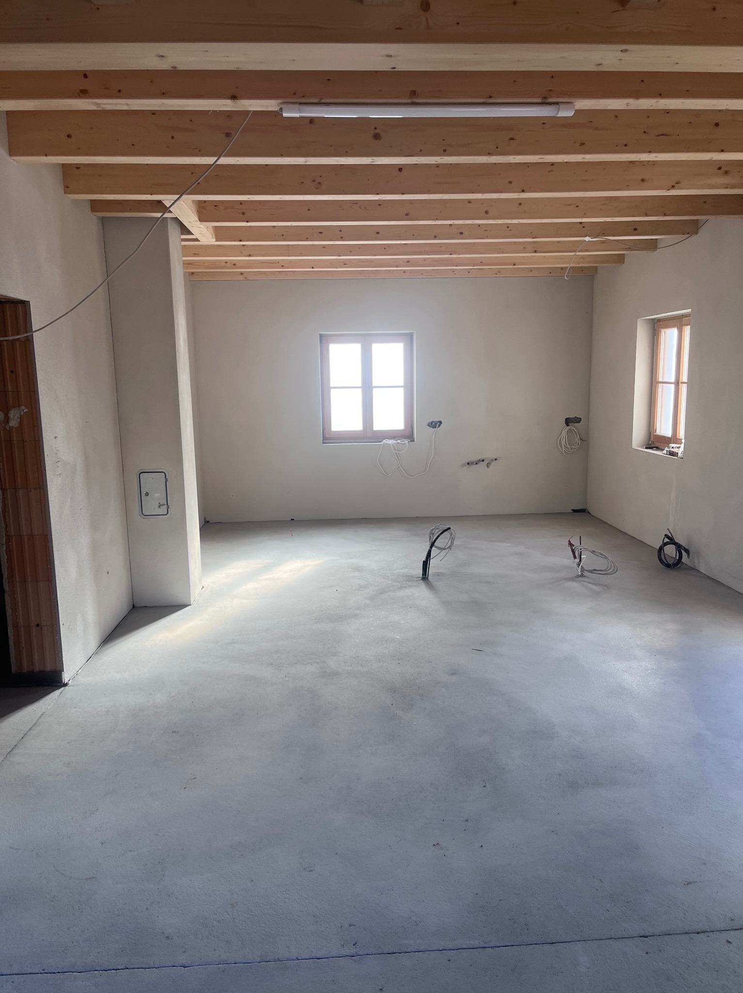 Surowe wnętrze z drewnianym stropem i świeżo wylaną posadzką. Widoczne kable instalacji elektrycznej, ściany przygotowane pod dalsze prace wykończeniowe. Dwa okna.