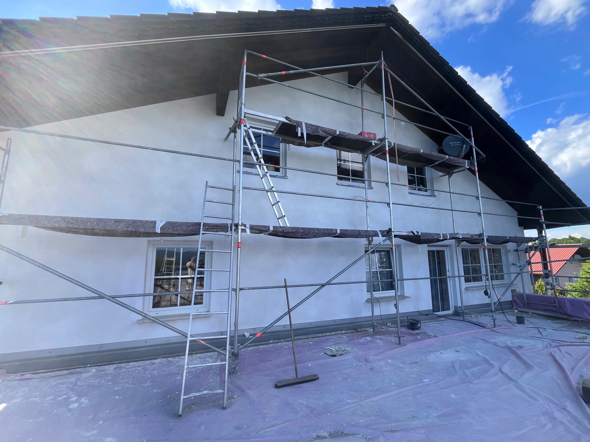 Dom jednorodzinny w trakcie prac elewacyjnych, widoczne rusztowanie, świeżo nałożony tynk i zabezpieczona folią przestrzeń robocza.