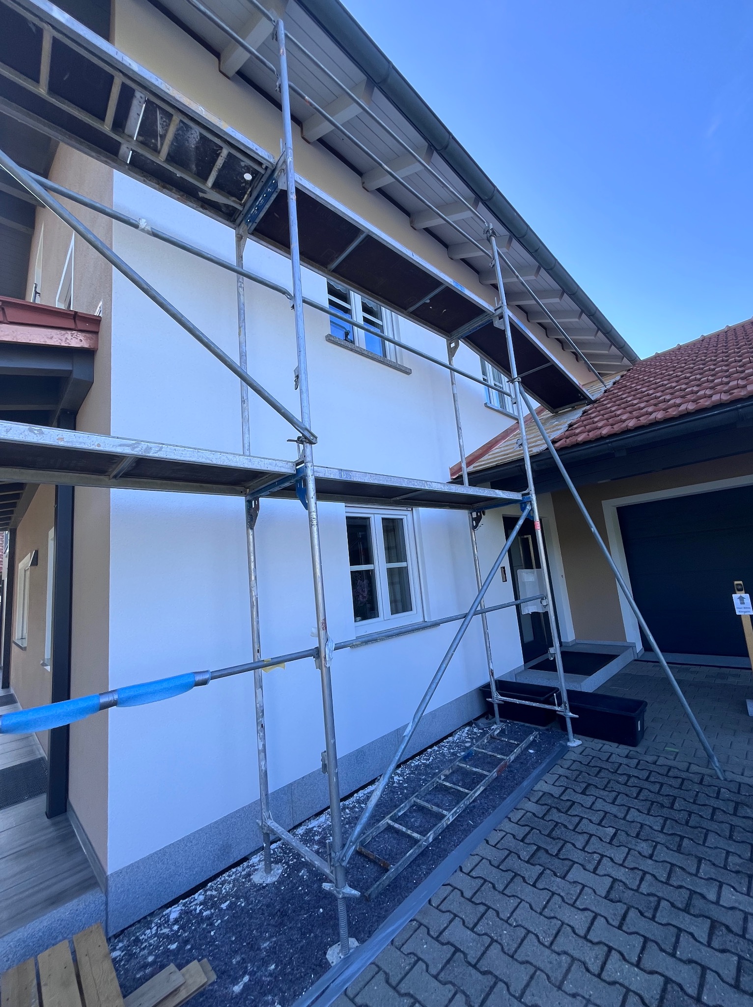 Dom jednorodzinny w rusztowaniach po świeżym, białym tynkowaniu. Widoczne okna, dachówka i fragment chodnika z kostki. Niebieskie niebo w tle.