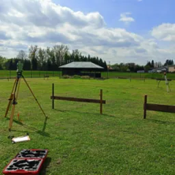 Prace pomiarowe w terenie: Dwa statywy z niwelatorami na zielonej łące, widoczne drewniane konstrukcje, skrzynka narzędziowa. Błękitne niebo z chmurami w tle.