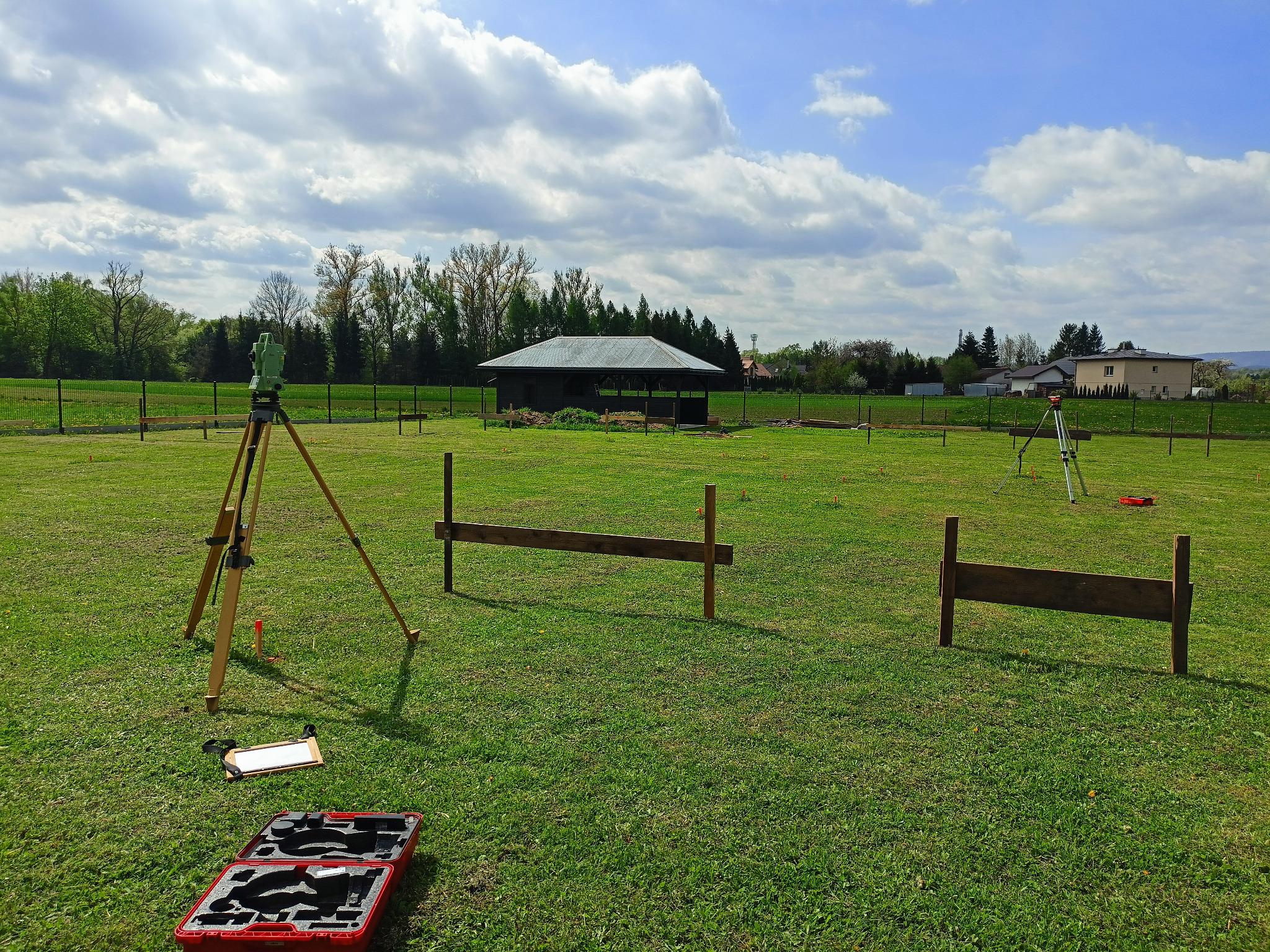 Prace pomiarowe w terenie: Dwa statywy z niwelatorami na zielonej łące, widoczne drewniane konstrukcje, skrzynka narzędziowa. Błękitne niebo z chmurami w tle.