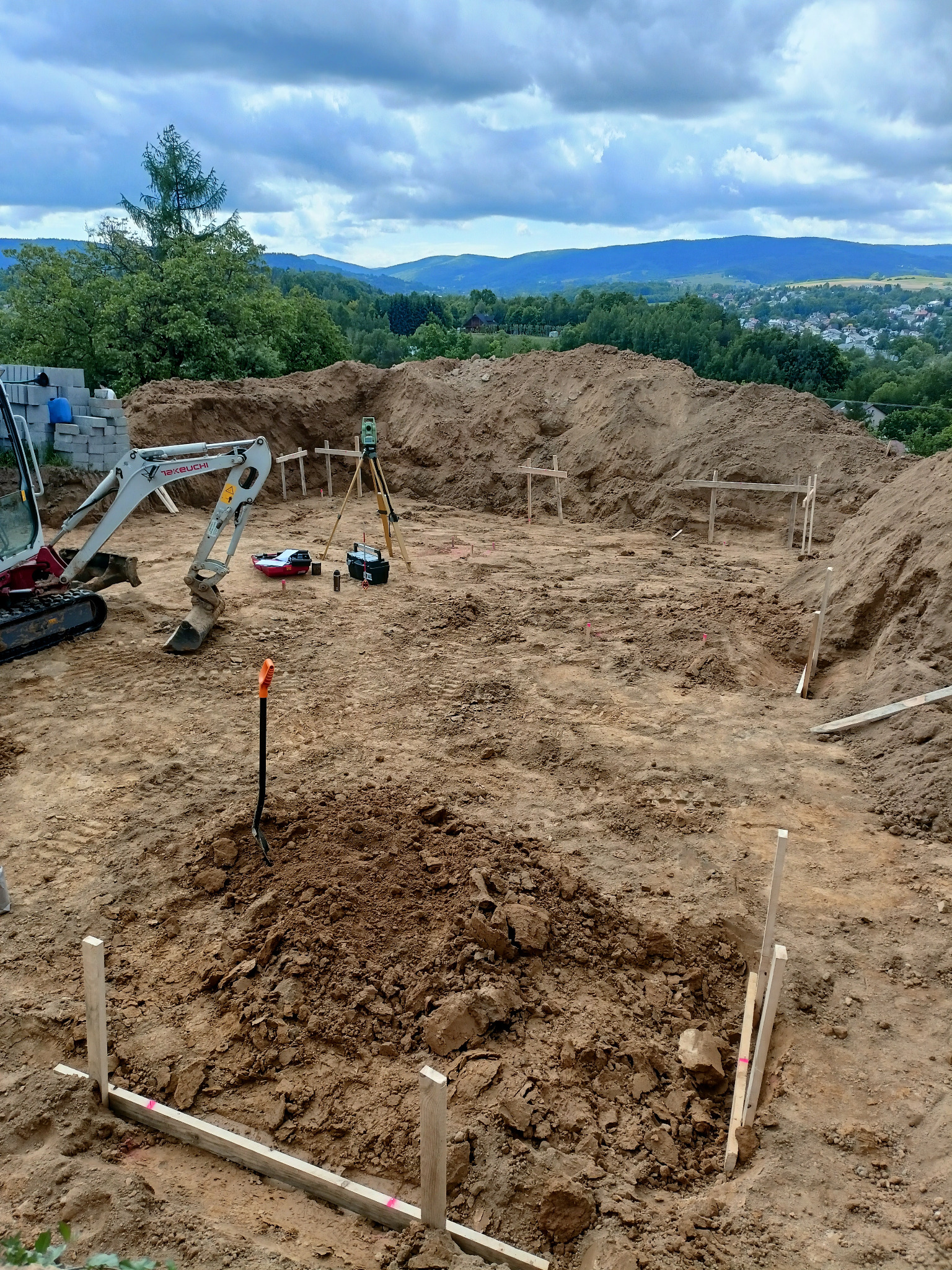 Wytyczenie fundamentów pod budowę domu z użyciem niwelatora, palików i małej koparki na działce z widokiem na góry.