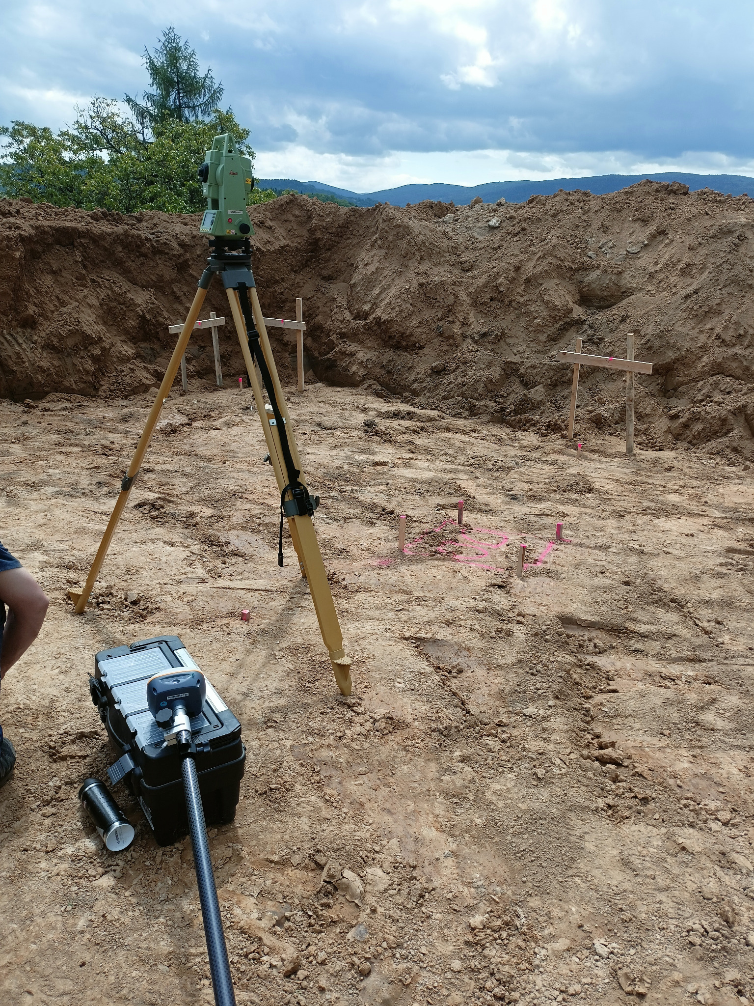 Zielony teodolit na trójnogu na placu budowy, wyznaczający punkty pomiarowe zaznaczone palikami i różową farbą. W tle wzgórza i pochmurne niebo.
