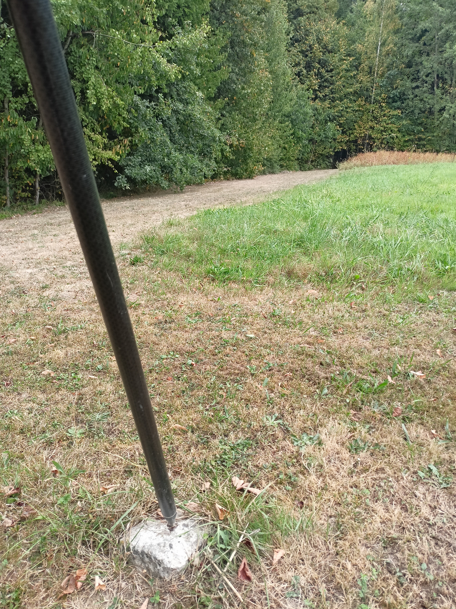 Czarny tyczka geodezyjna wbita w betonowy słupek na zewnątrz, z polną drogą i drzewami w tle. Pomiar terenu.