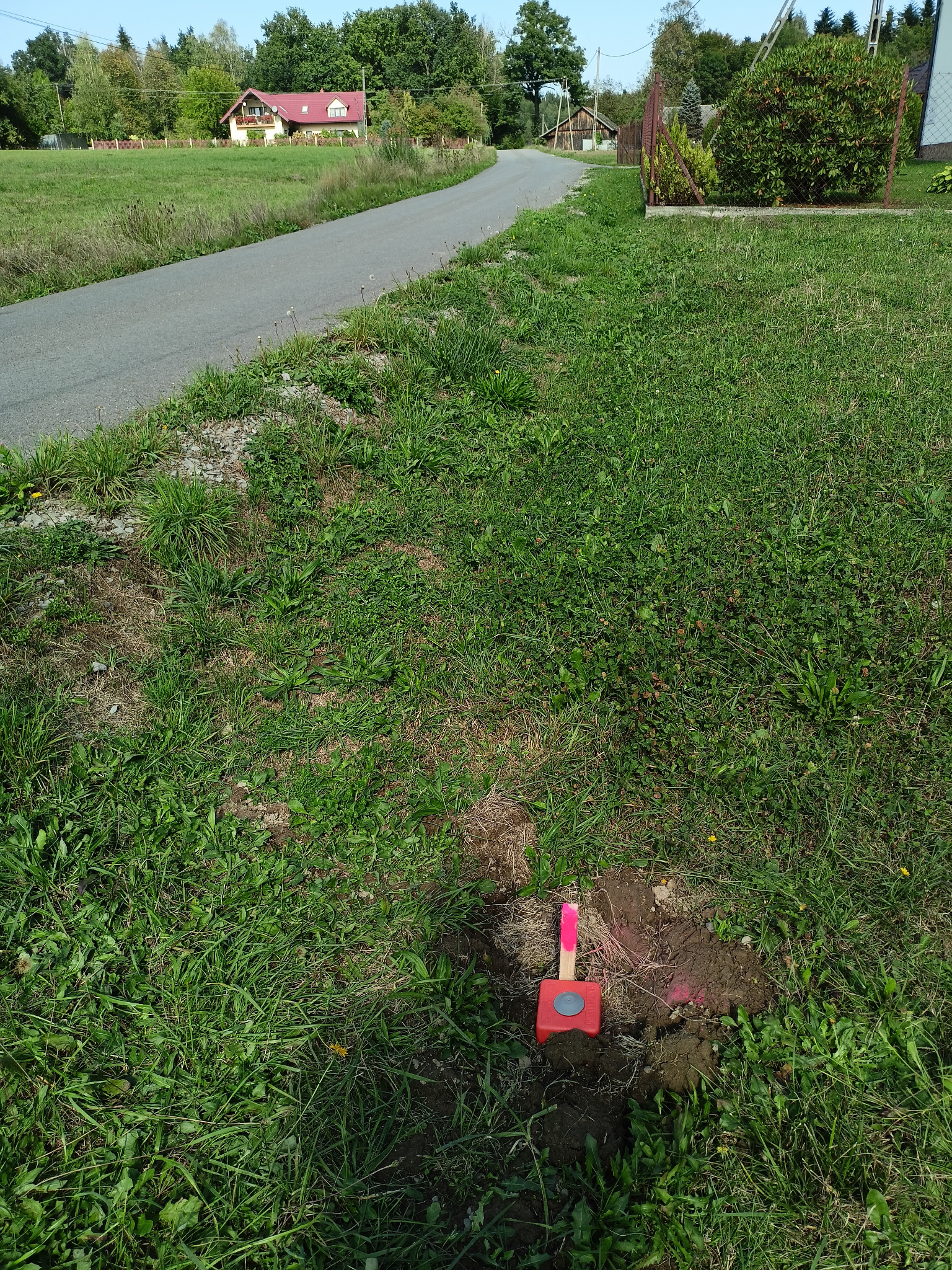 Czerwony palik geodezyjny wbity w ziemię na tle zielonej trawy i wiejskiej drogi. W oddali widoczny dom.