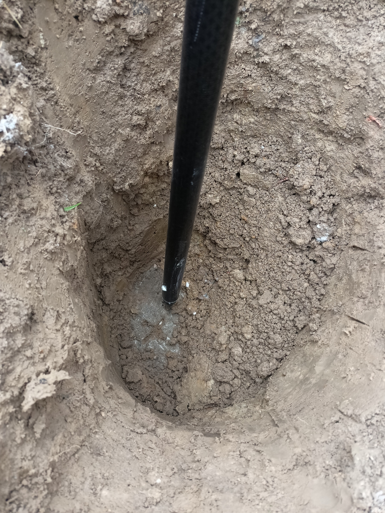 Czarny pręt wbity w wykop ziemny, prawdopodobnie podczas prac pomiarowych. Widoczna struktura gleby i ślady po wbiciu.