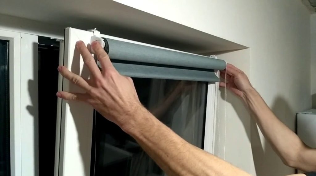 Montaż rolety okiennej w białej ramie. Widoczne ręce montera regulujące mechanizm. Szary materiał rolety częściowo zasłania okno.