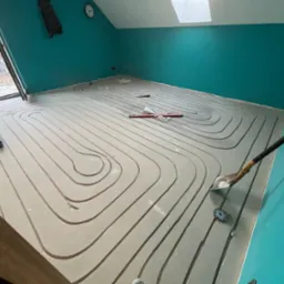 Wnętrze poddasza z turkusowymi ścianami w trakcie montażu ogrzewania podłogowego. Widoczne ułożone rury grzewcze, narzędzia pracy i okno dachowe.