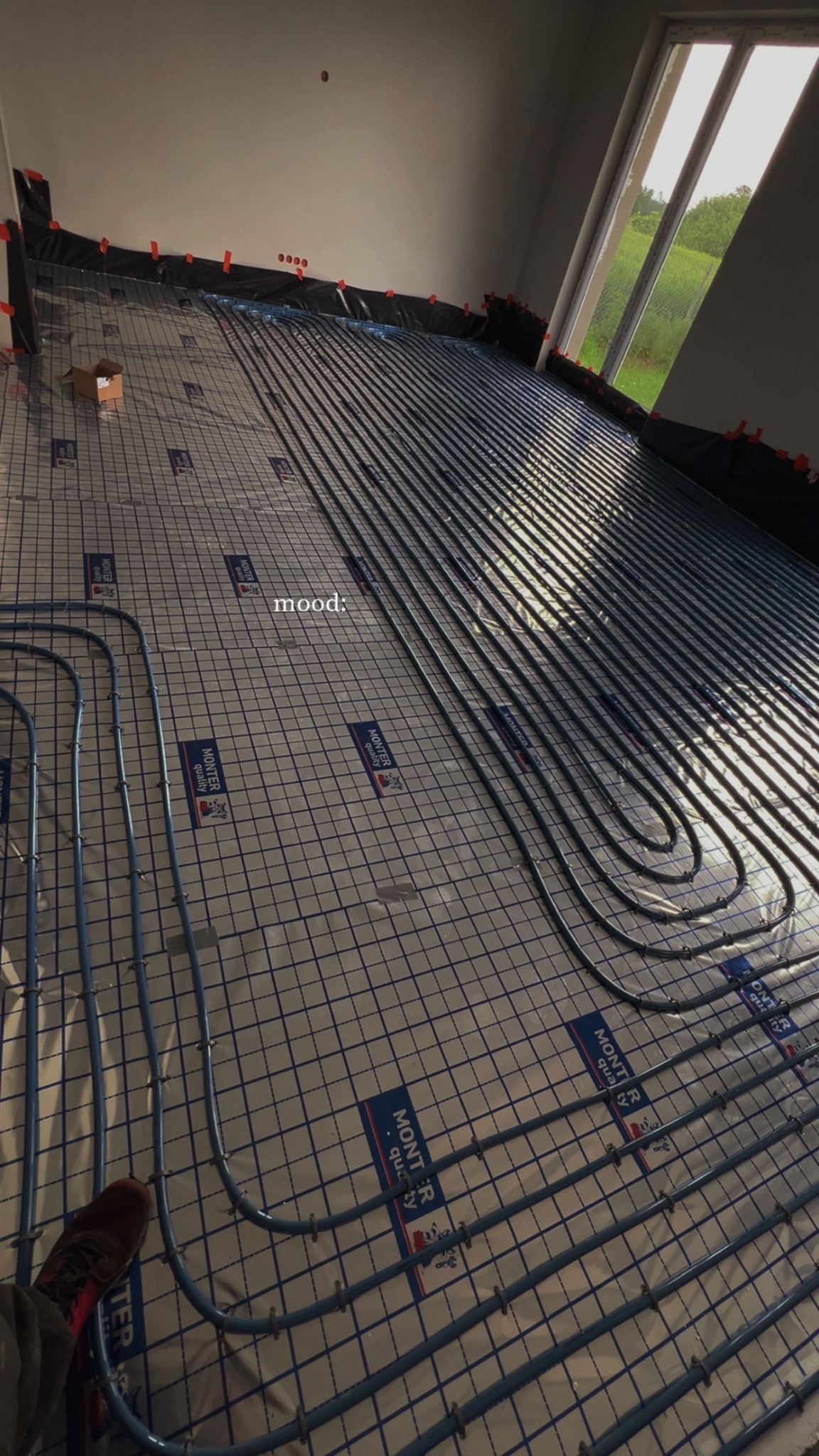 Niebieskie rury ogrzewania podłogowego ułożone na srebrnej macie z siatką w jasnym wnętrzu. Widoczne mocowania, fragment okna i but. Etap instalacji przed zalaniem posadzką.