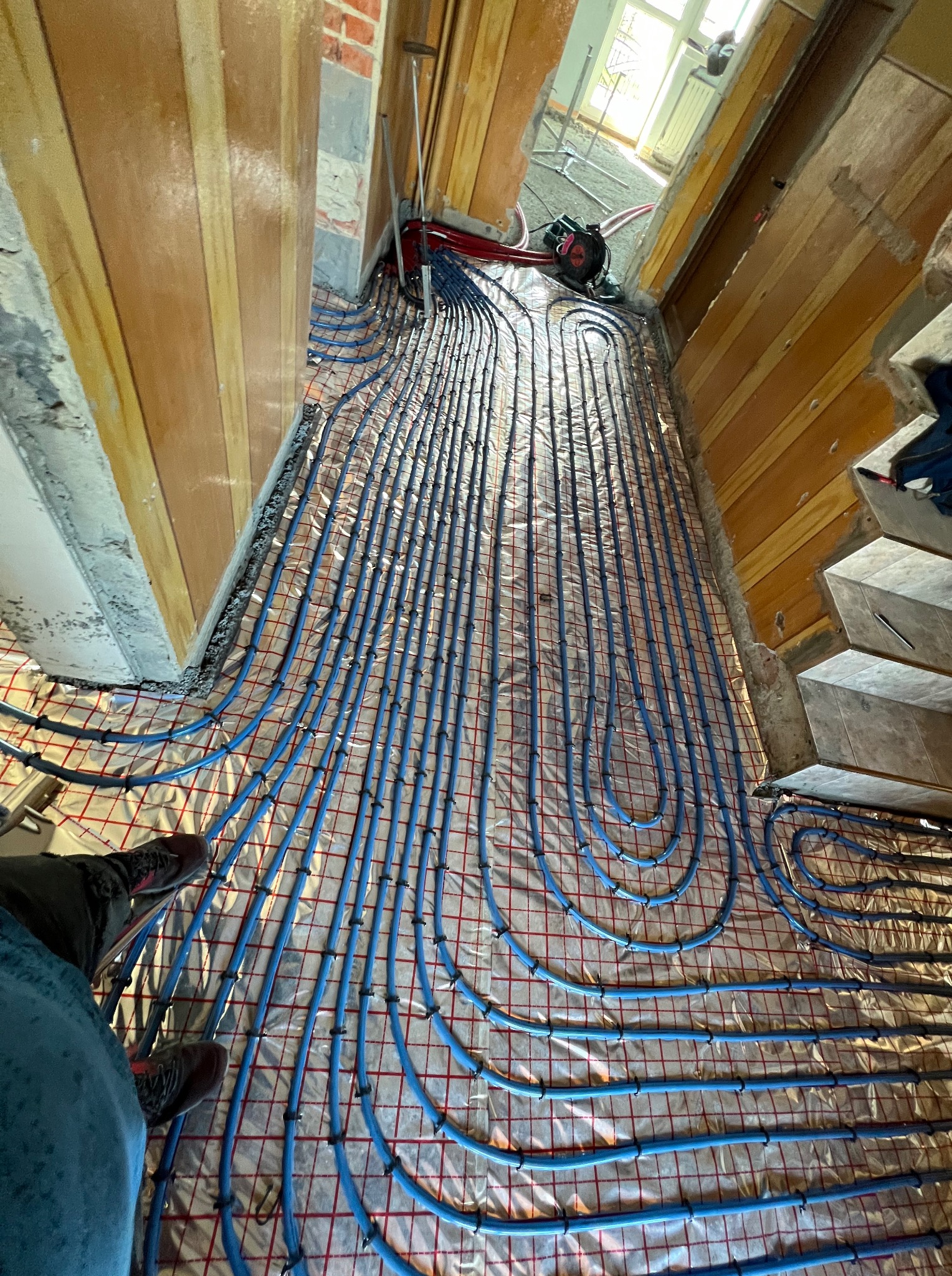 Instalacja ogrzewania podłogowego z niebieskich rur na folii z siatką, widok z góry na etapie montażu w nowym budynku. Fragment nogi w kadrze.