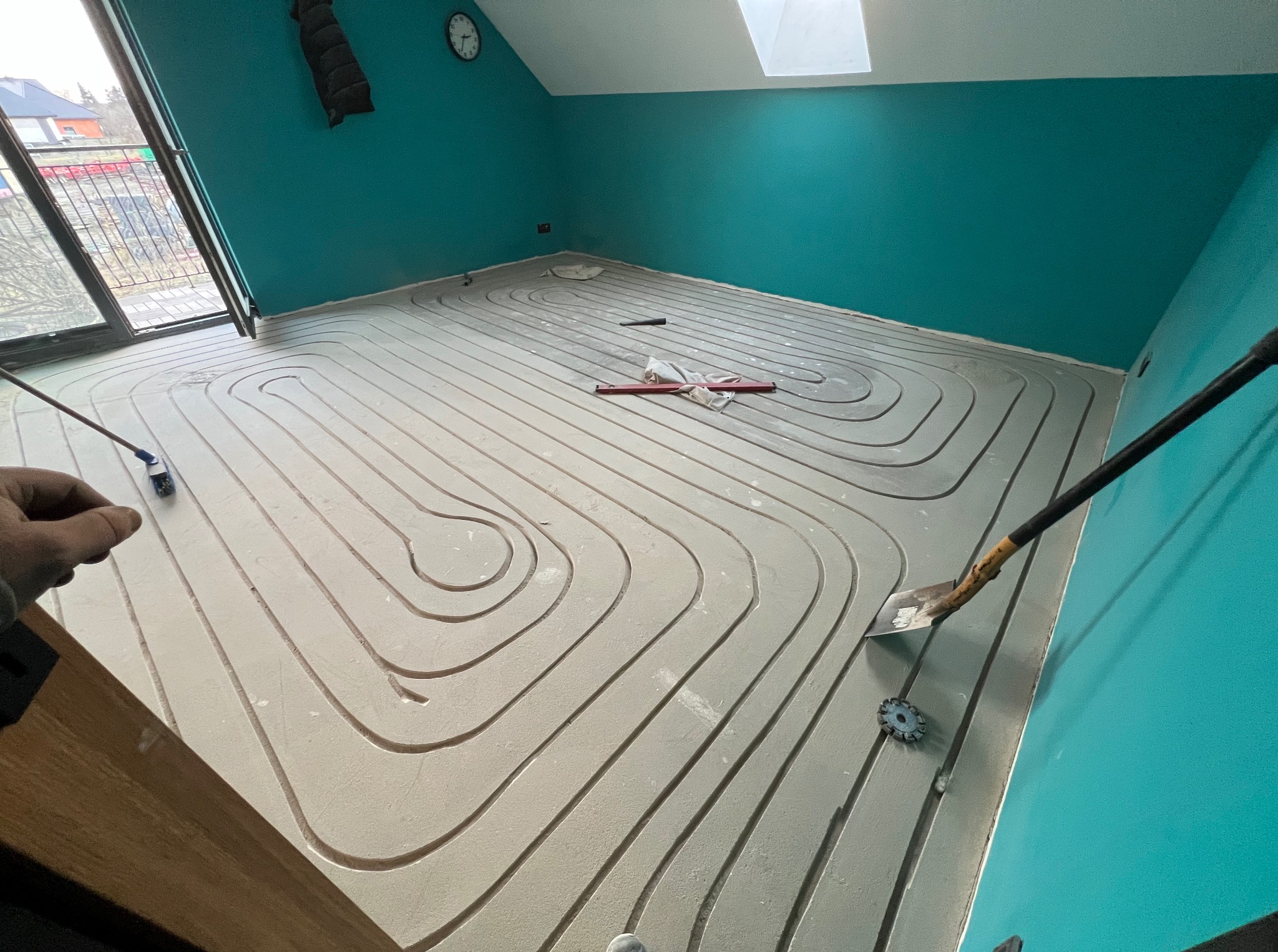 Wnętrze poddasza z turkusowymi ścianami w trakcie montażu ogrzewania podłogowego. Widoczne ułożone rury grzewcze, narzędzia pracy i okno dachowe.