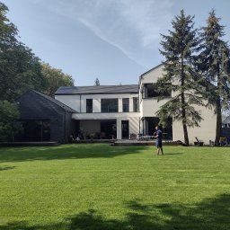 AGAPANT - Rozległy trawnik przed nowoczesnym domem, mężczyzna podlewa trawnik w słoneczny dzień. Widoczne drzewa i budynki w tle. Trawa świeżo skoszona, równa.