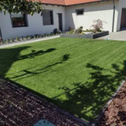 Idealnie równy trawnik z rolki przed nowoczesnym domem. Ciemne cienie drzew na zielonej trawie. Estetyczne obrzeża i rabaty z korą.