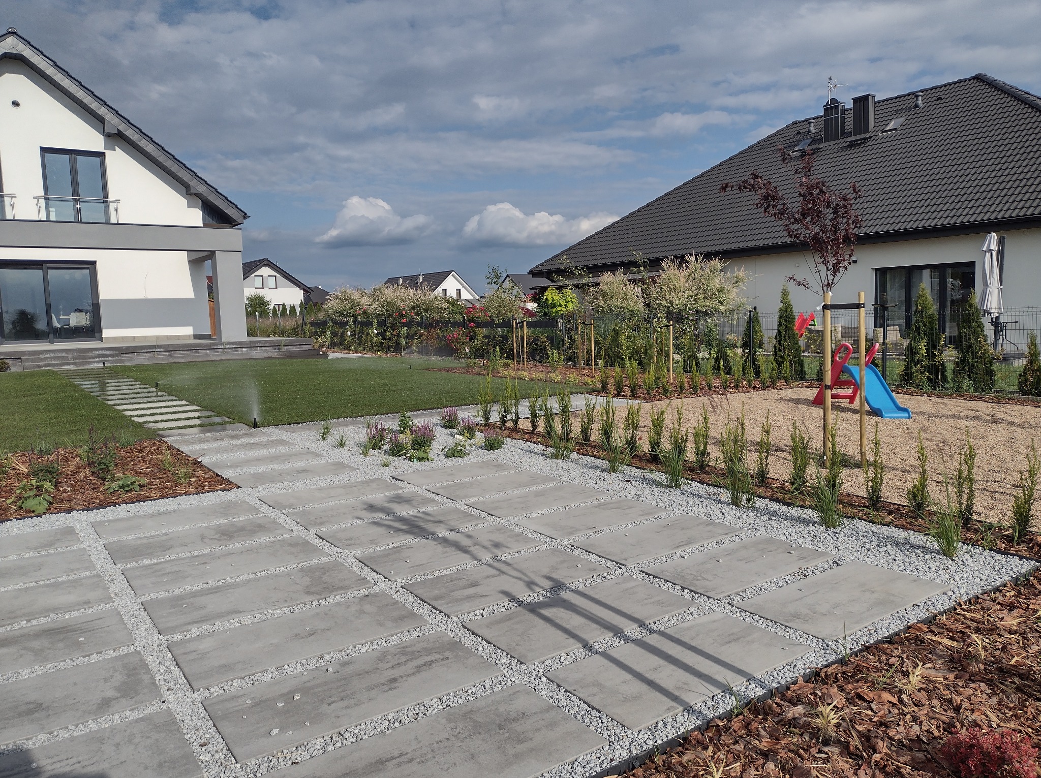 Nowoczesny ogród przydomowy z betonową ścieżką wysypaną drobnym żwirem, rabatami z roślinami i placem zabaw dla dzieci na żwirowej nawierzchni.