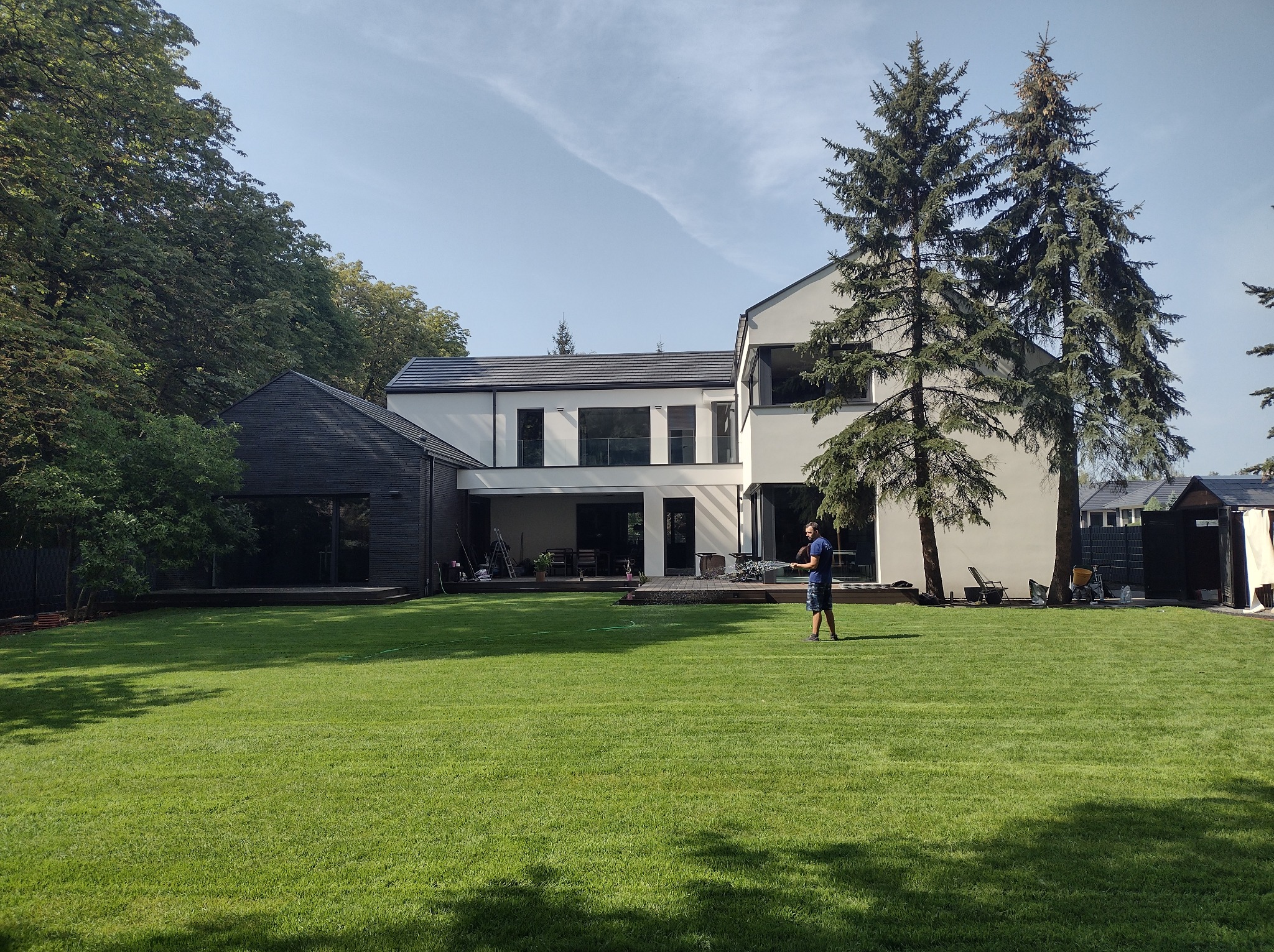 Rozległy trawnik przed nowoczesnym domem, mężczyzna podlewa trawnik w słoneczny dzień. Widoczne drzewa i budynki w tle. Trawa świeżo skoszona, równa.
