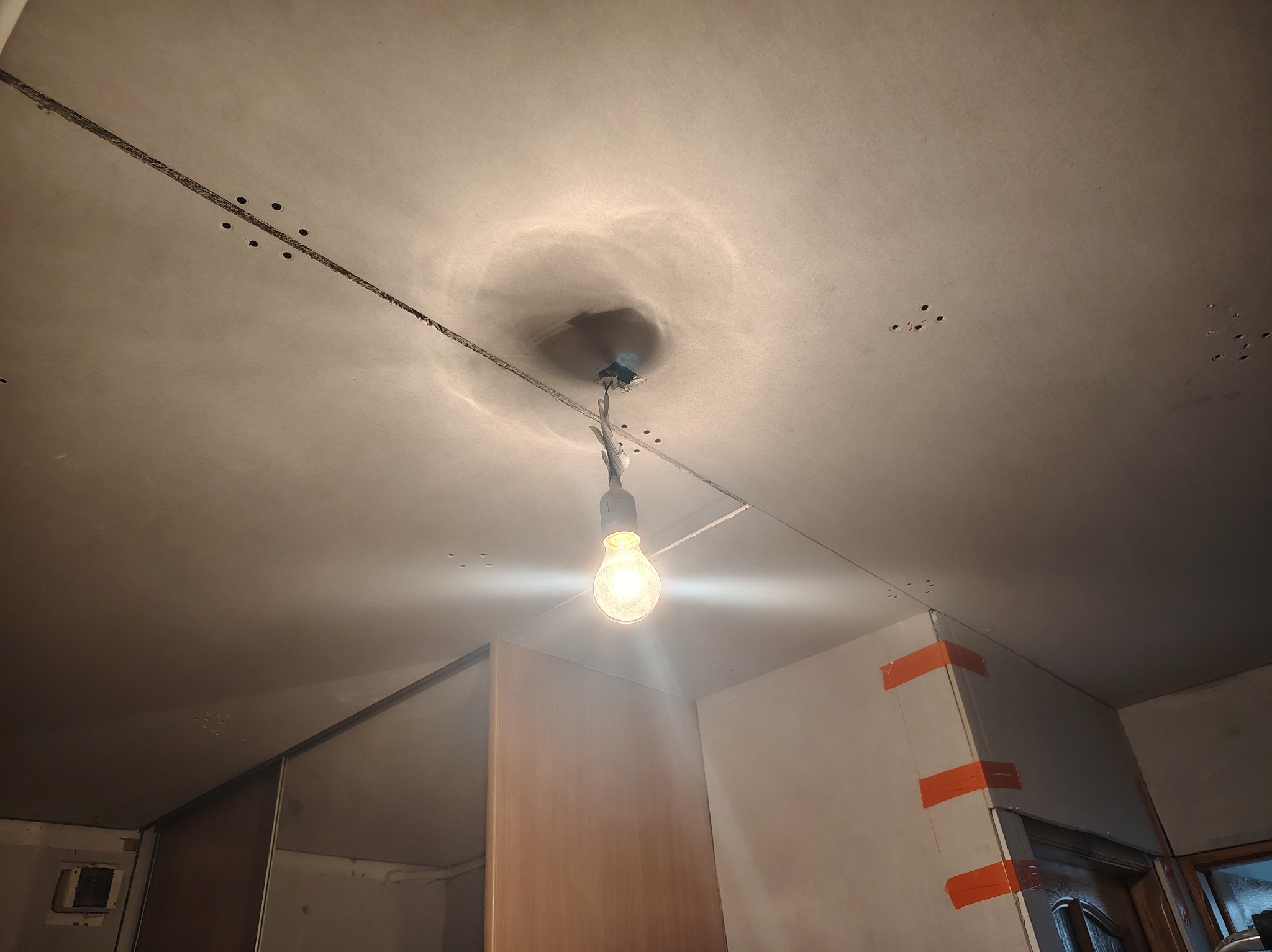 Montaż sufitu podwieszanego z płyt karton-gips: widoczne łączenia, otwory po wkrętach i prowizoryczne oświetlenie żarówką. Etap prac wykończeniowych.