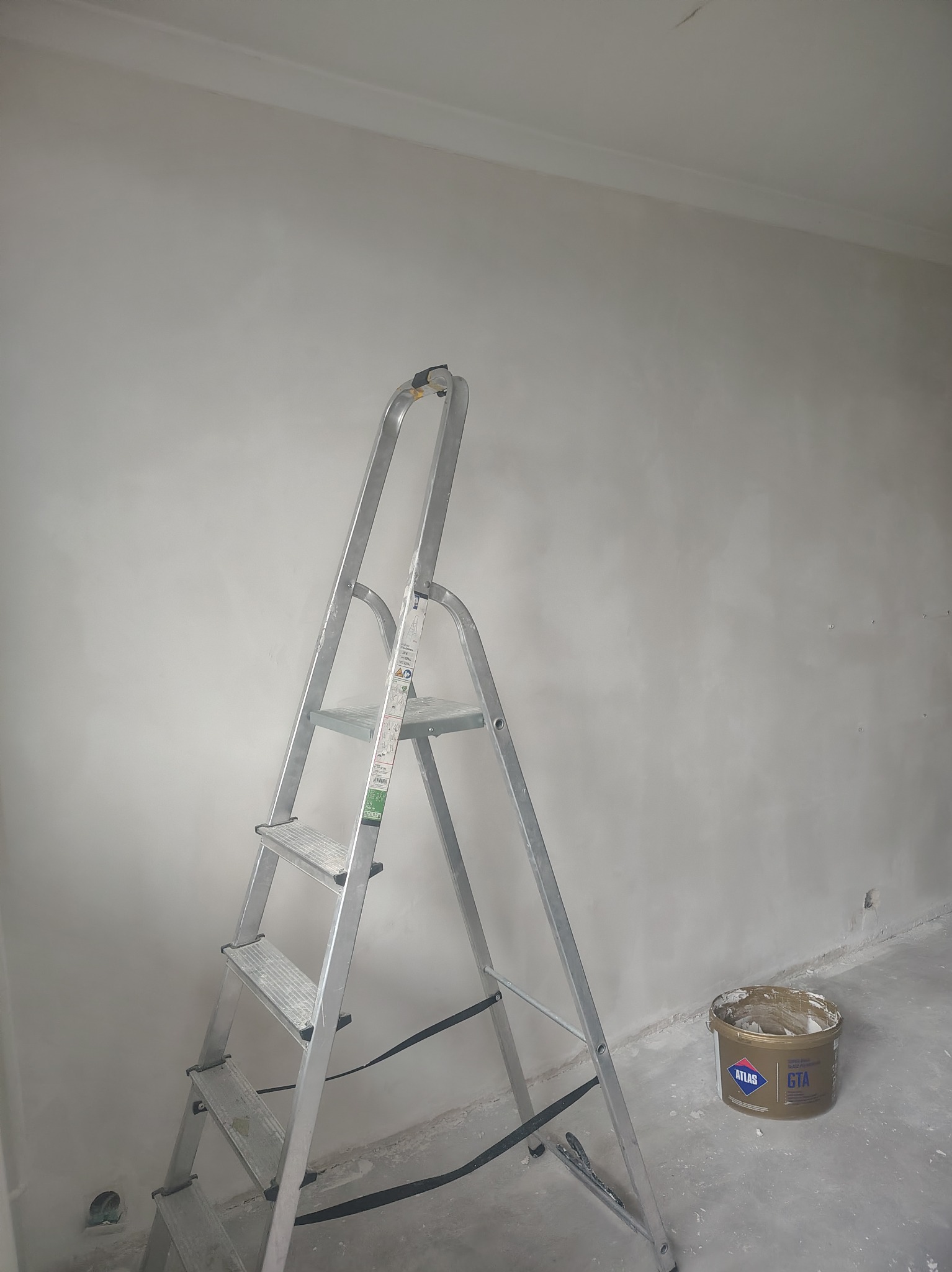 Ściana po szpachlowaniu z widocznymi śladami, drabina aluminiowa i wiadro po szpachli Atlas GTA. Przygotowanie do malowania w trakcie remontu.