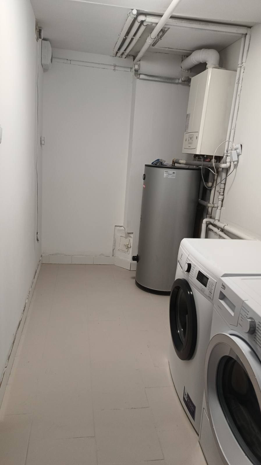 Białe pomieszczenie gospodarcze z piecem gazowym, bojlerem i pralką. Widoczne rury pod sufitem i płytki na podłodze. Ściany pomalowane na biało.