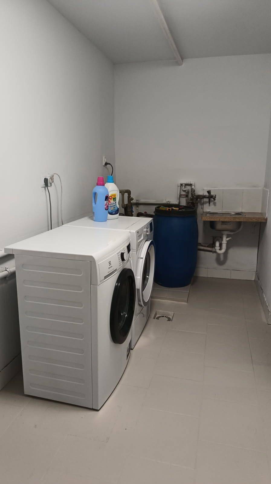 Biała pralka i suszarka w pomieszczeniu gospodarczym z niebieską beczką i umywalką. Na pralce stoją środki czystości. Jasne płytki na podłodze.