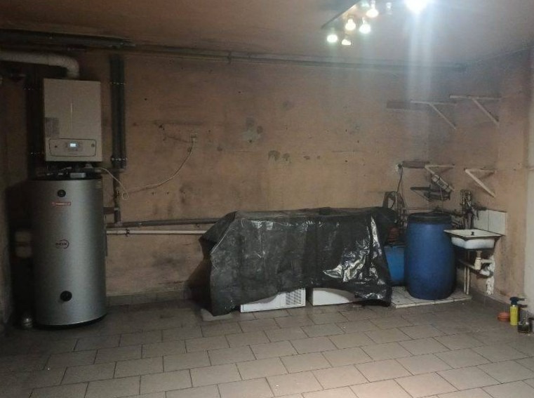 Piwnica z piecem CO, bojlerem, niebieską beczką i zlewem. Podłoga wyłożona płytkami, ściany w odcieniach beżu. Na suficie widoczne oświetlenie.