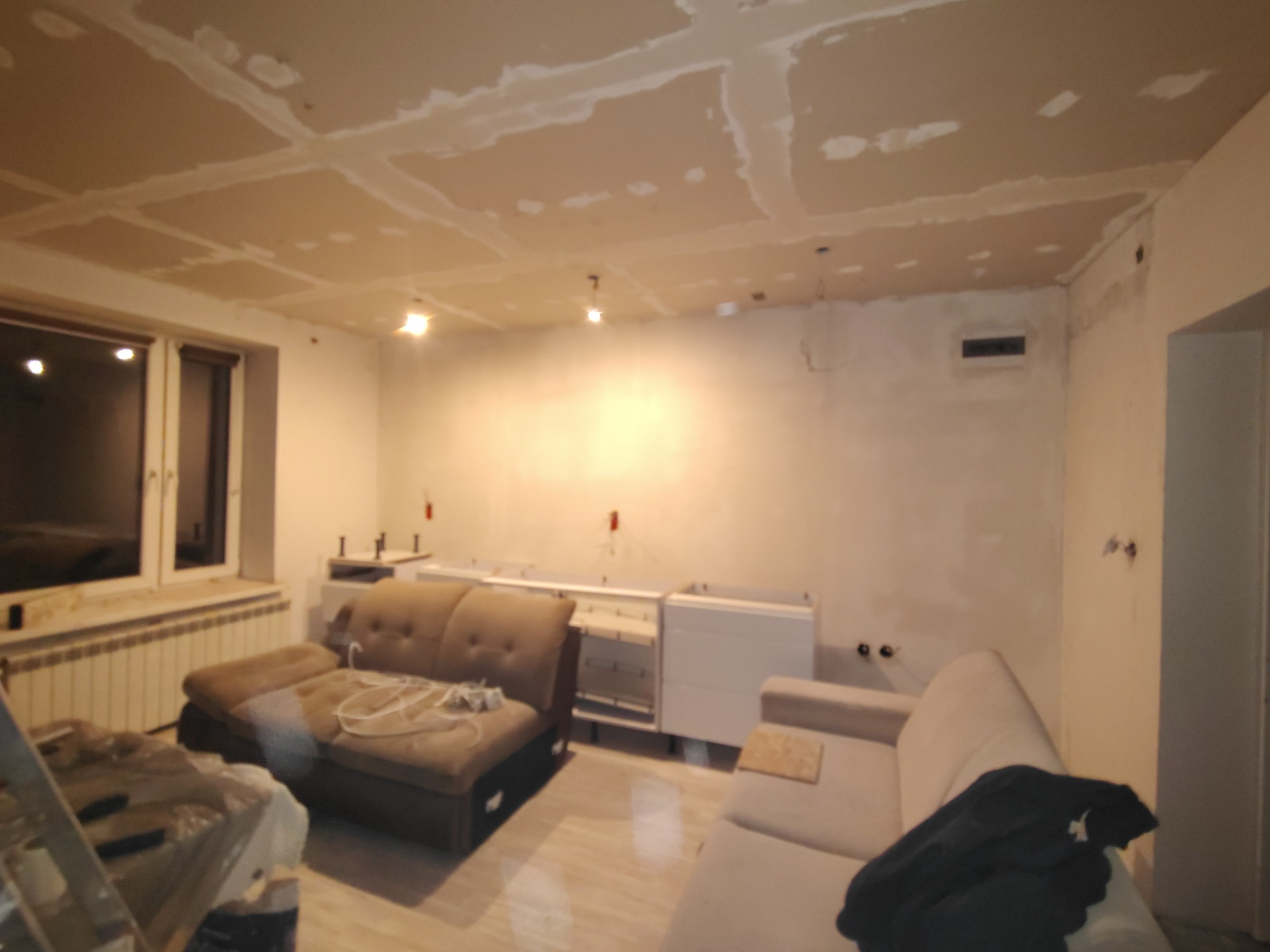 Wnętrze w trakcie remontu: szpachlowana sufit z płyt karton-gips, przygotowane ściany, częściowo zamontowane meble kuchenne, sofy w pokoju. Widok na remontowany pokój.