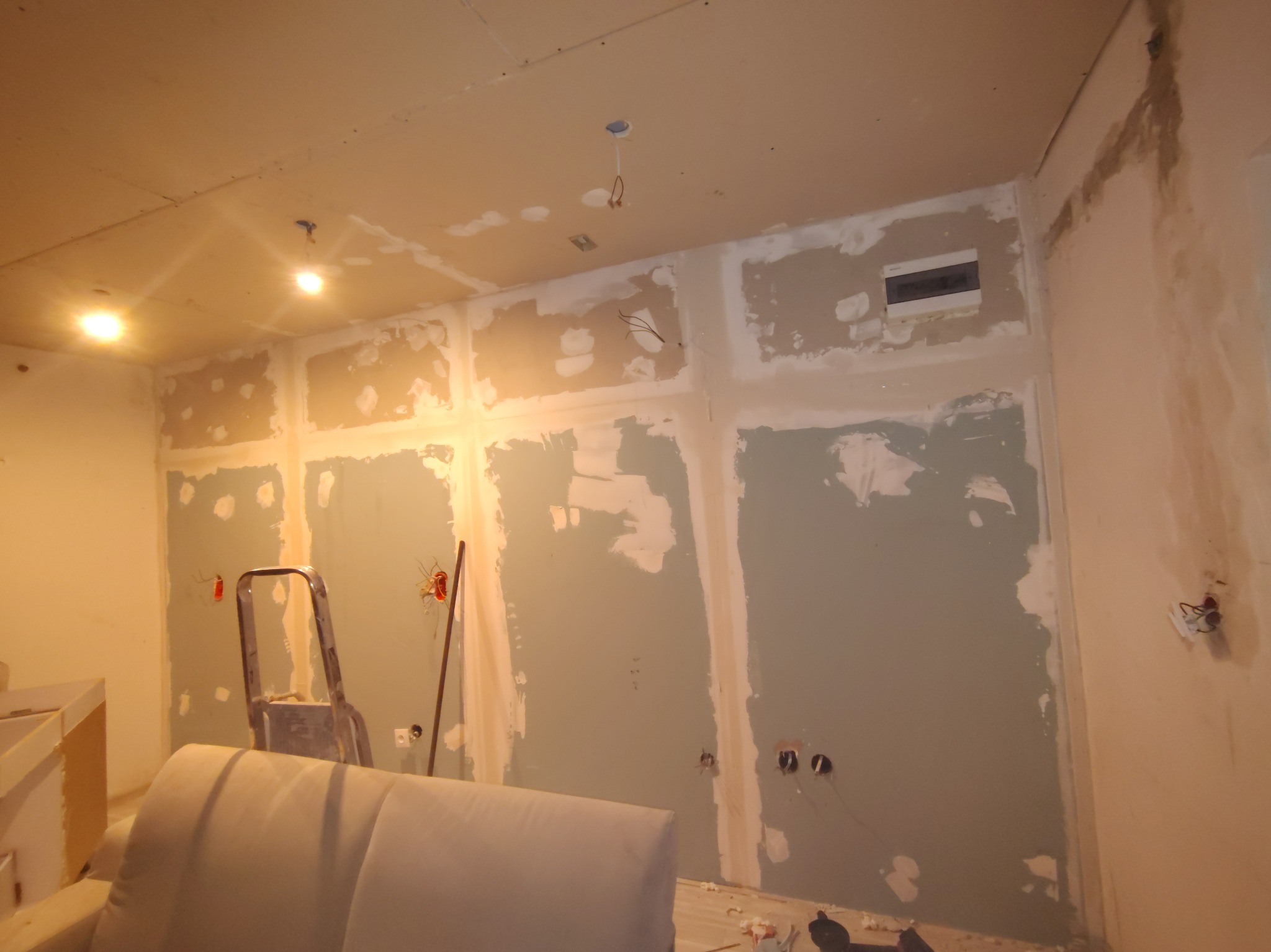 Wnętrze w trakcie szpachlowania: ściany z widocznymi punktami elektrycznymi, drabina i fragment mebla. Prace wykończeniowe w pomieszczeniu z surowym oświetleniem.