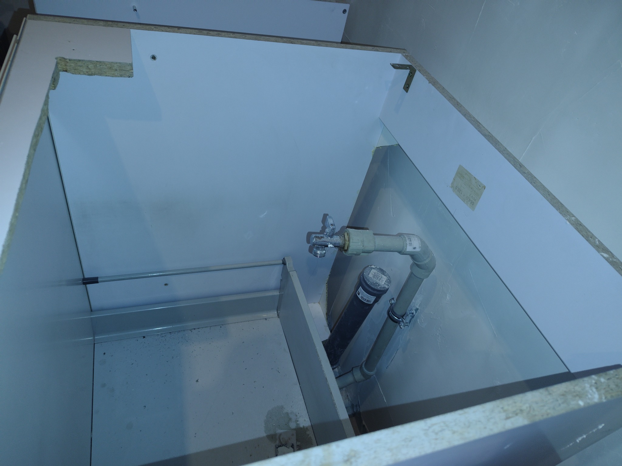 Wnętrze szafki pod zlew z widocznymi rurami i zaworem, wykonane z białych płyt meblowych. Widok z góry, ukazujący instalację hydrauliczną.