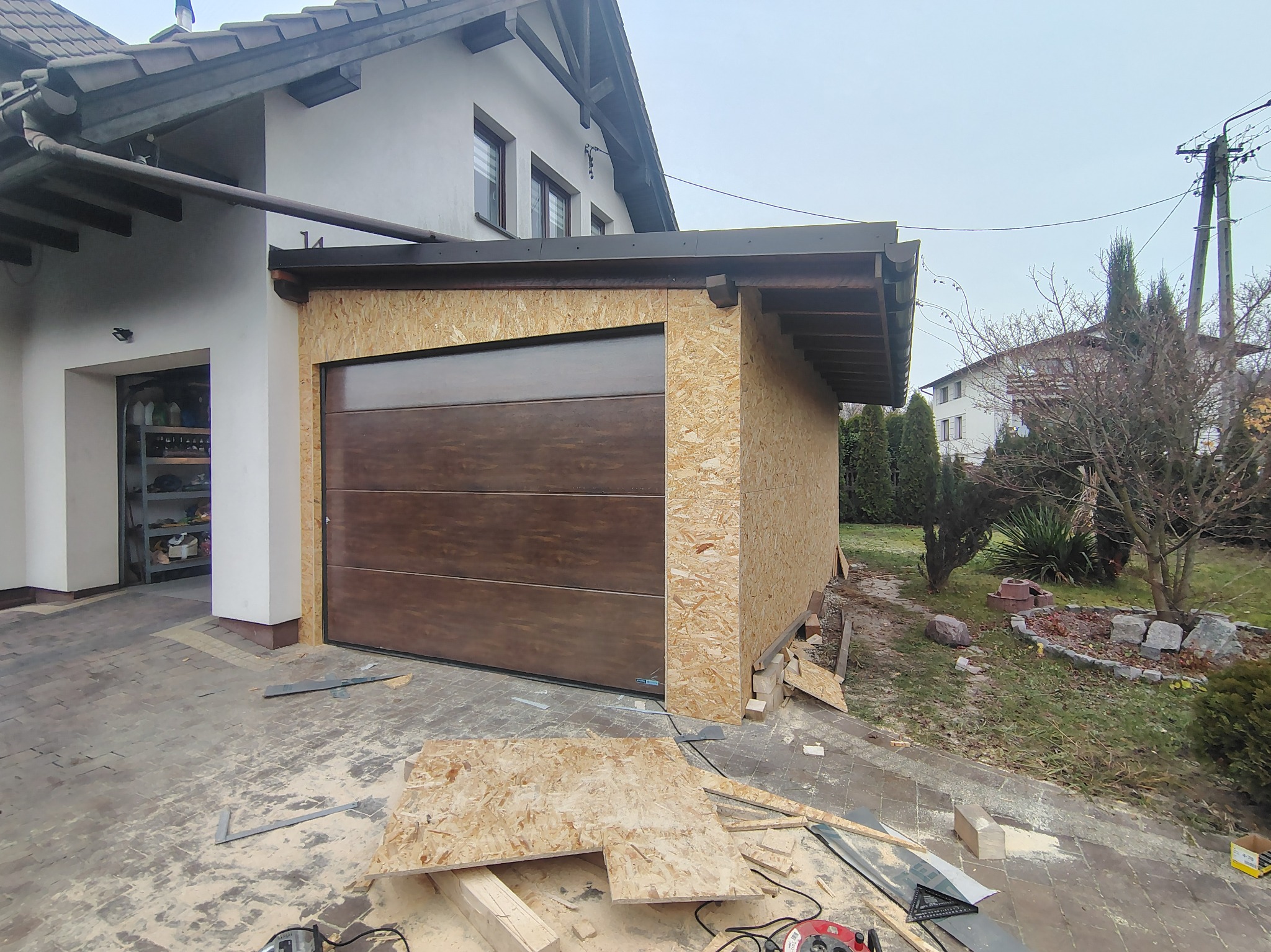 Montaż bramy garażowej w Porąbce: brązowa brama segmentowa w surowej obudowie z płyt OSB, widoczne narzędzia i materiały budowlane, dom w tle.