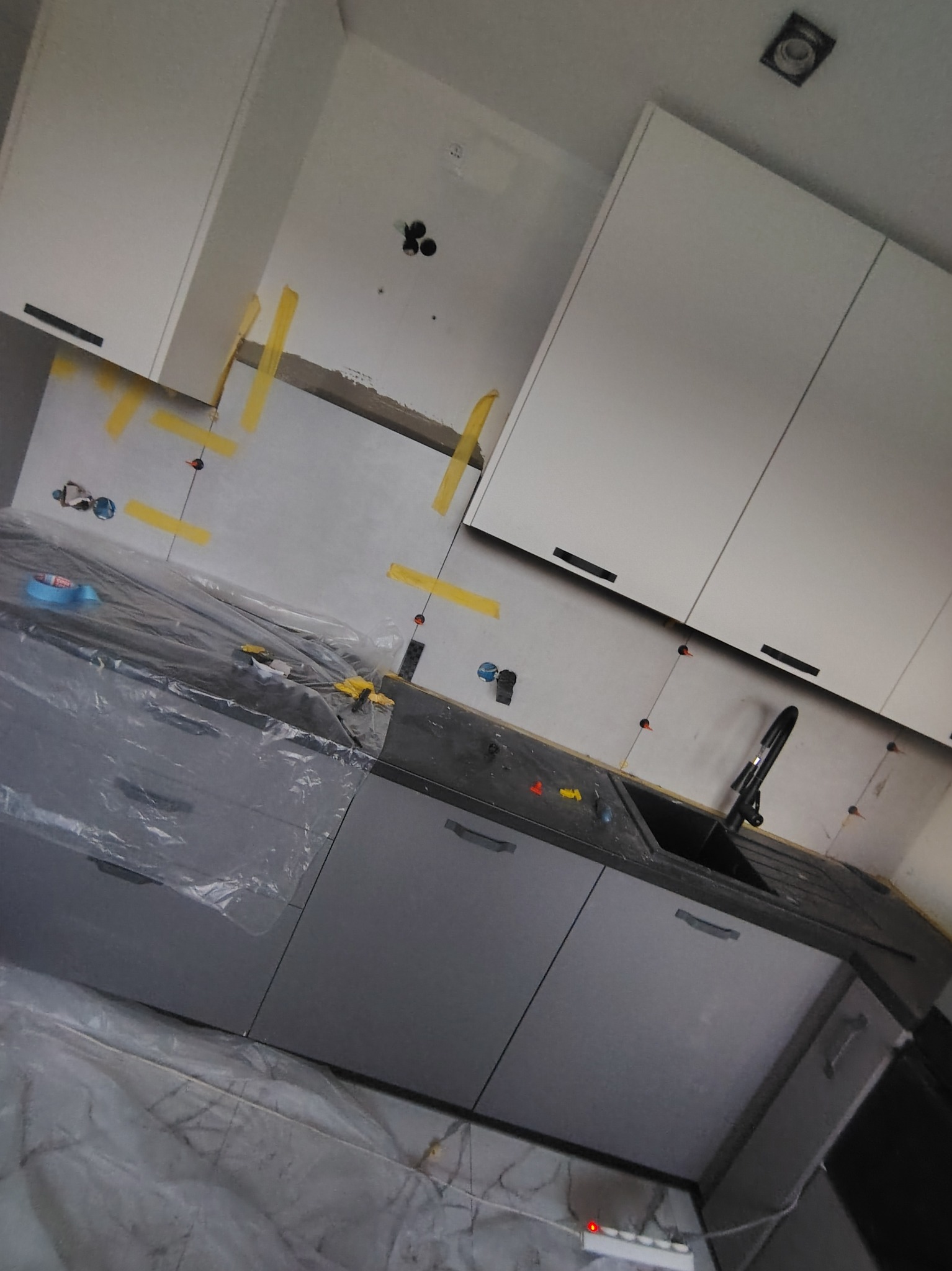 Montaż szarych szafek kuchennych z czarnymi uchwytami i baterią. Ściana z taśmą malarską, zabezpieczony blat folią. Widoczne punkty podłączeń.