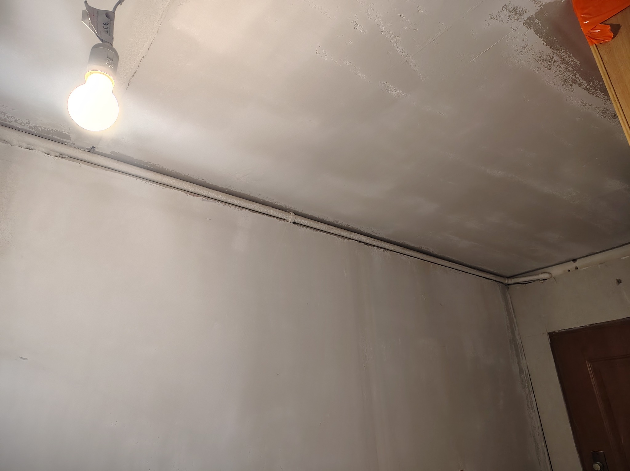 Szpachlowana ściana i sufit w trakcie prac wykończeniowych, widoczna żarówka i fragment drzwi. Przygotowanie do malowania.