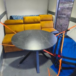 KUBA KAWALEC KKONCEPT - Wnętrze busa z zabezpieczonymi meblami: okrągły stół z blatem imitującym marmur, żółta sofa i niebieski koc, wszystko spięte pomarańczowymi pasami transportowymi.