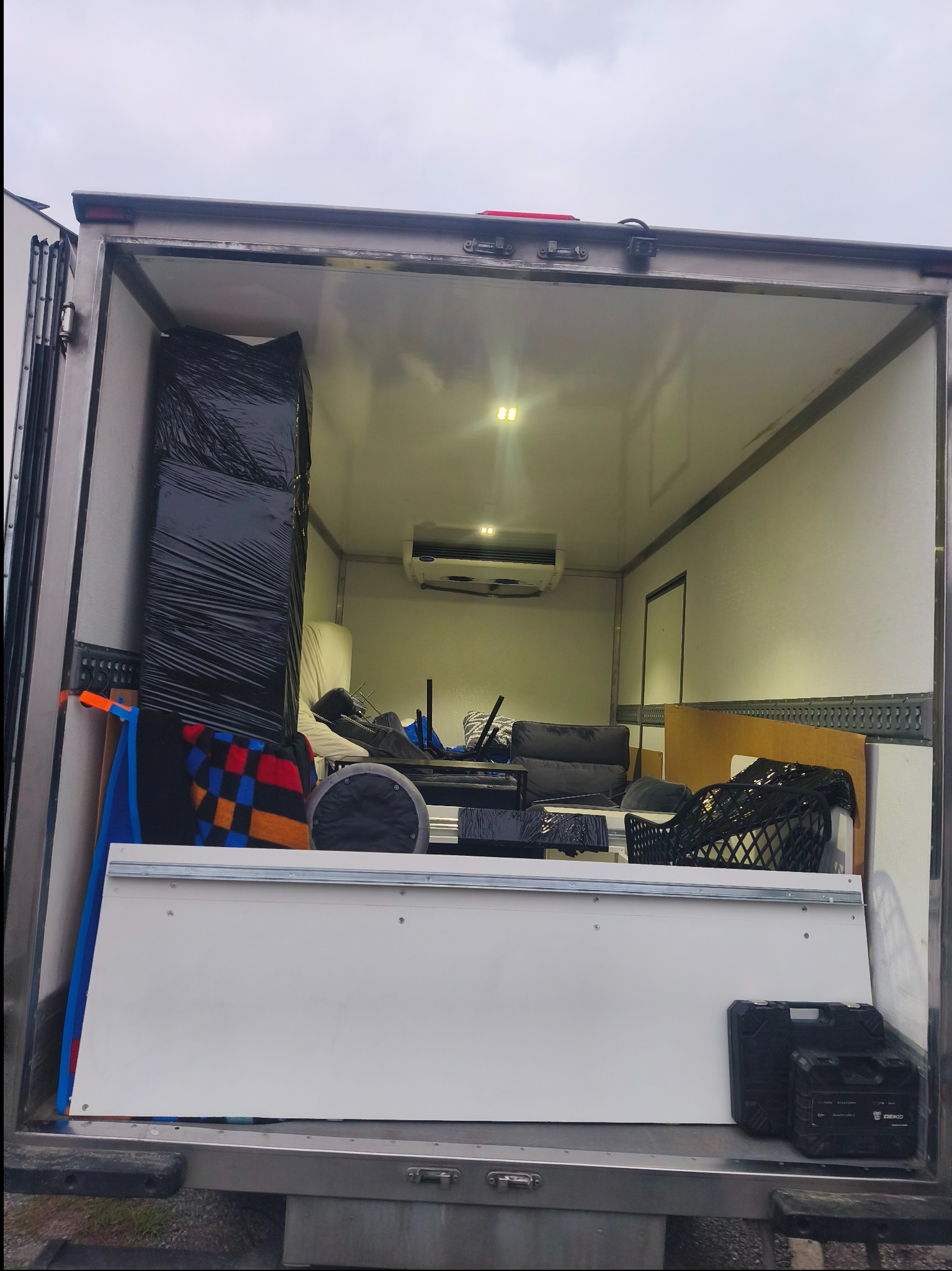 Wnętrze furgonetki załadowanej meblami i sprzętami, zabezpieczonymi folią i kocami. Widoczny system chłodzenia w górnej części przestrzeni ładunkowej.