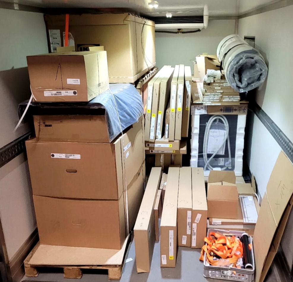 Wnętrze chłodni wypełnione kartonami, meblami w paczkach i zabezpieczeniami ładunku. Towar przygotowany do transportu, częściowo na palecie.