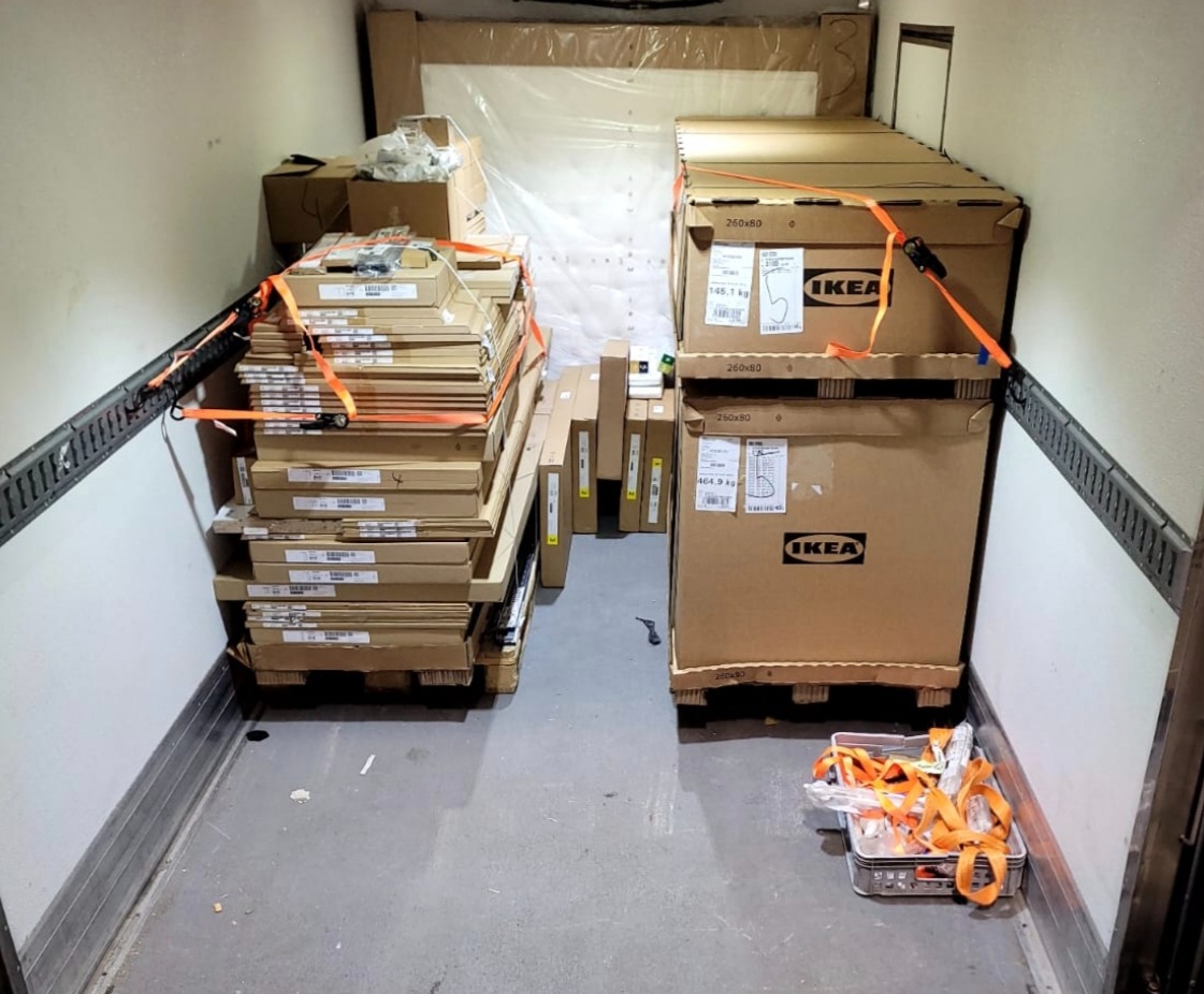 Wnętrze busa wypełnione kartonami IKEA, zabezpieczonymi pomarańczowymi pasami transportowymi, gotowe do przewozu. Widoczne palety i różnorodne rozmiary paczek.