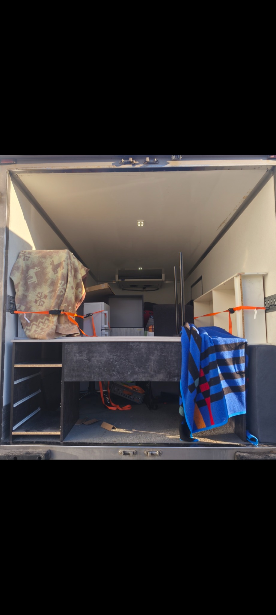 Wnętrze busa załadowane meblami i sprzętami, zabezpieczone pasami transportowymi i kocami. Widok z tyłu pojazdu, wnętrze oświetlone.